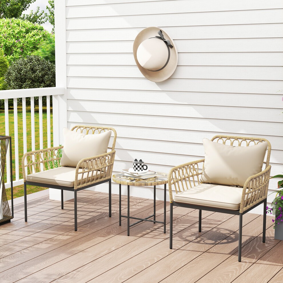 Three-piece rattan bistro set featuring two beige cushioned chairs and a glass-top table, set against a white wall and greenery.