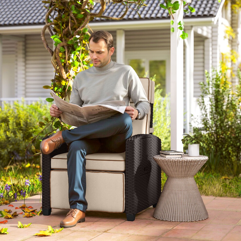 Dark brown rattan garden recliner sun lounger with a flip table, featuring a cushioned seat and backrest, set in a green outdoor space with a person reading a newspaper.