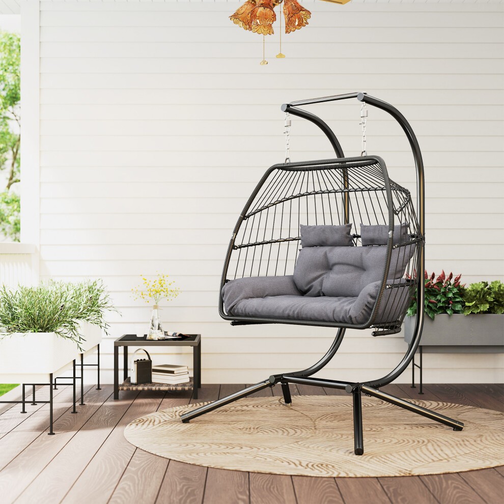 Rattan double hanging egg chair in dark grey with plush grey cushions, featuring a sturdy black metal stand, set on a wooden deck with potted plants in the background.