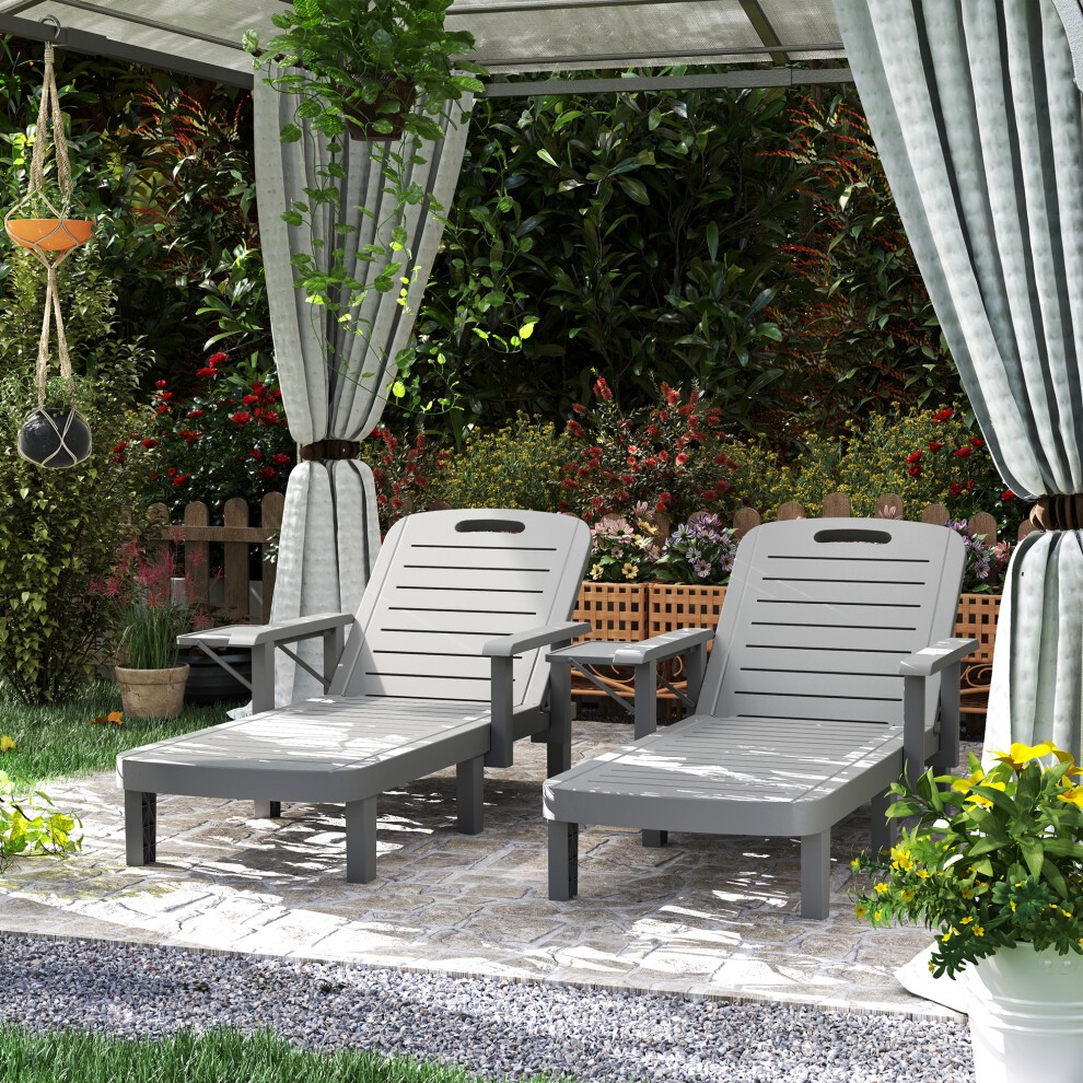 Set of 2 dark grey resin reclining sun loungers with slatted design, positioned under a canopy in a garden setting, surrounded by greenery and flowers.