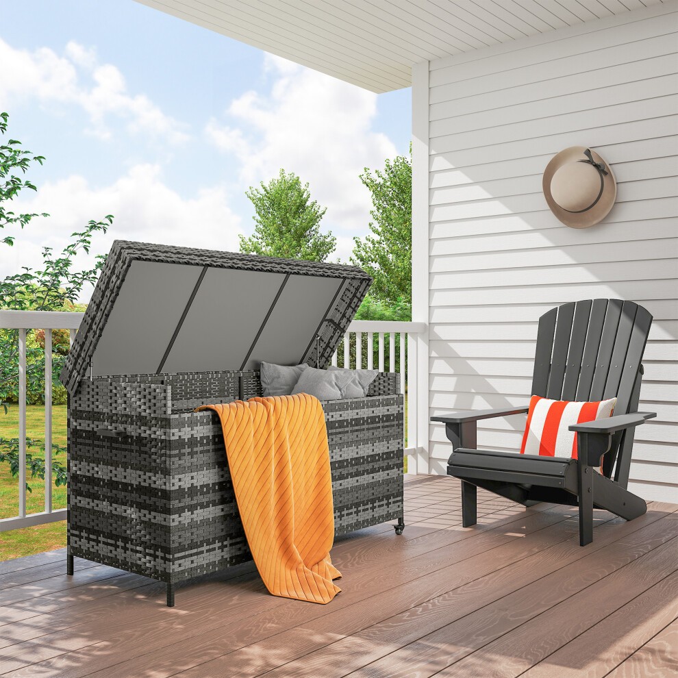 Grey rattan garden storage box with a gas lift lid, wheels for mobility, and a cozy blanket draped over it, placed on a wooden deck beside an Adirondack chair.