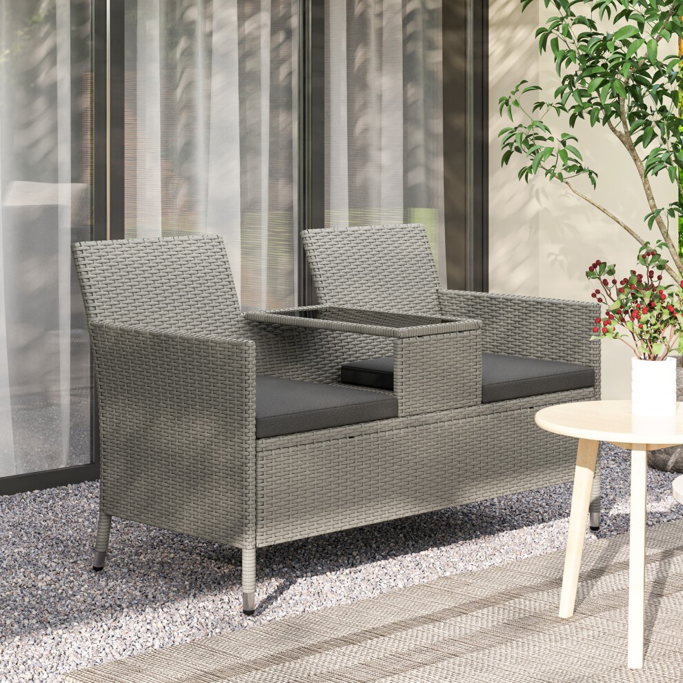 Grey rattan love seat with a central table, featuring cushioned seats and a sleek design, set against a backdrop of glass and greenery.