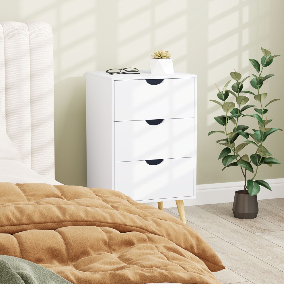 Cream white chest of drawers with three drawers, featuring black handles, wooden legs, placed beside a bed with a brown blanket and a potted plant in a bedroom setting.