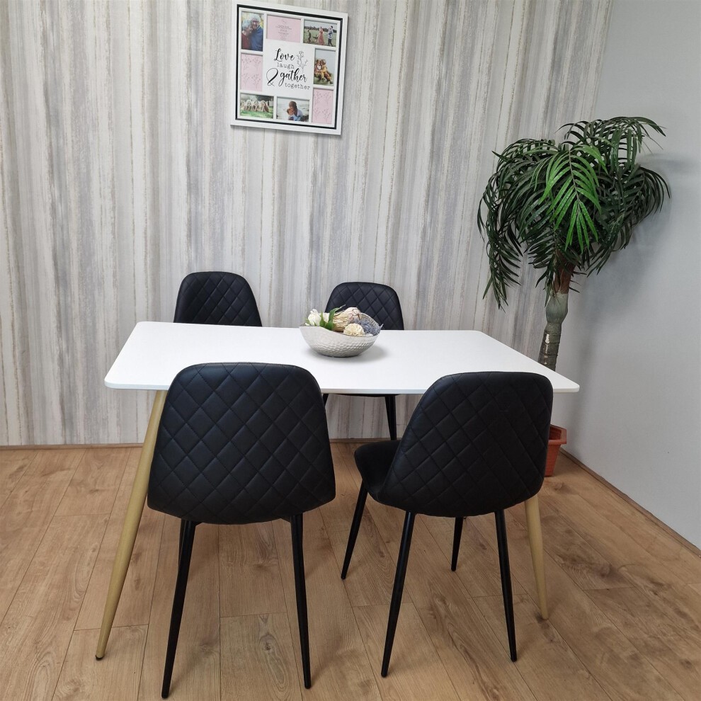 KOSY KOALA wooden white dining table with four black faux leather padded chairs featuring diamond stitching, set against a light wood wall and a potted plant.