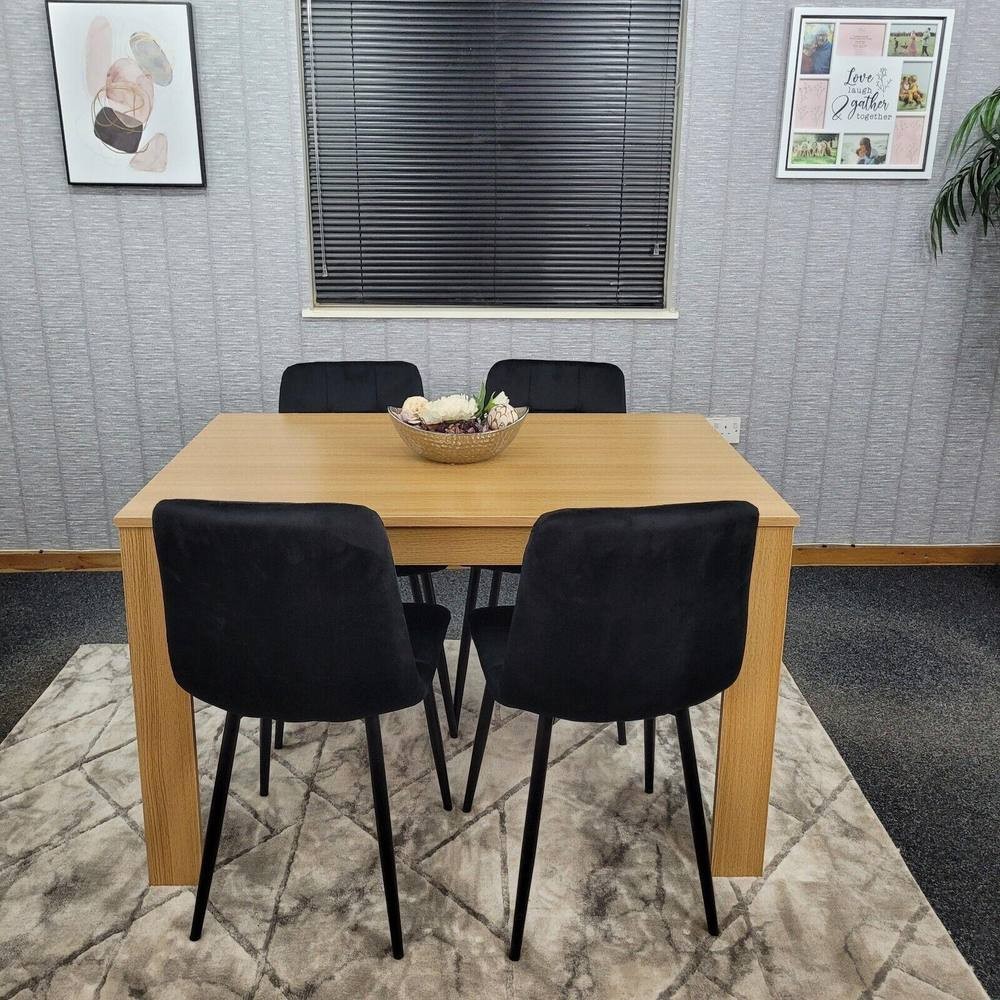 Beige wooden dining table set with four black upholstered chairs, featuring a central decorative bowl, set against a textured wall and patterned rug.