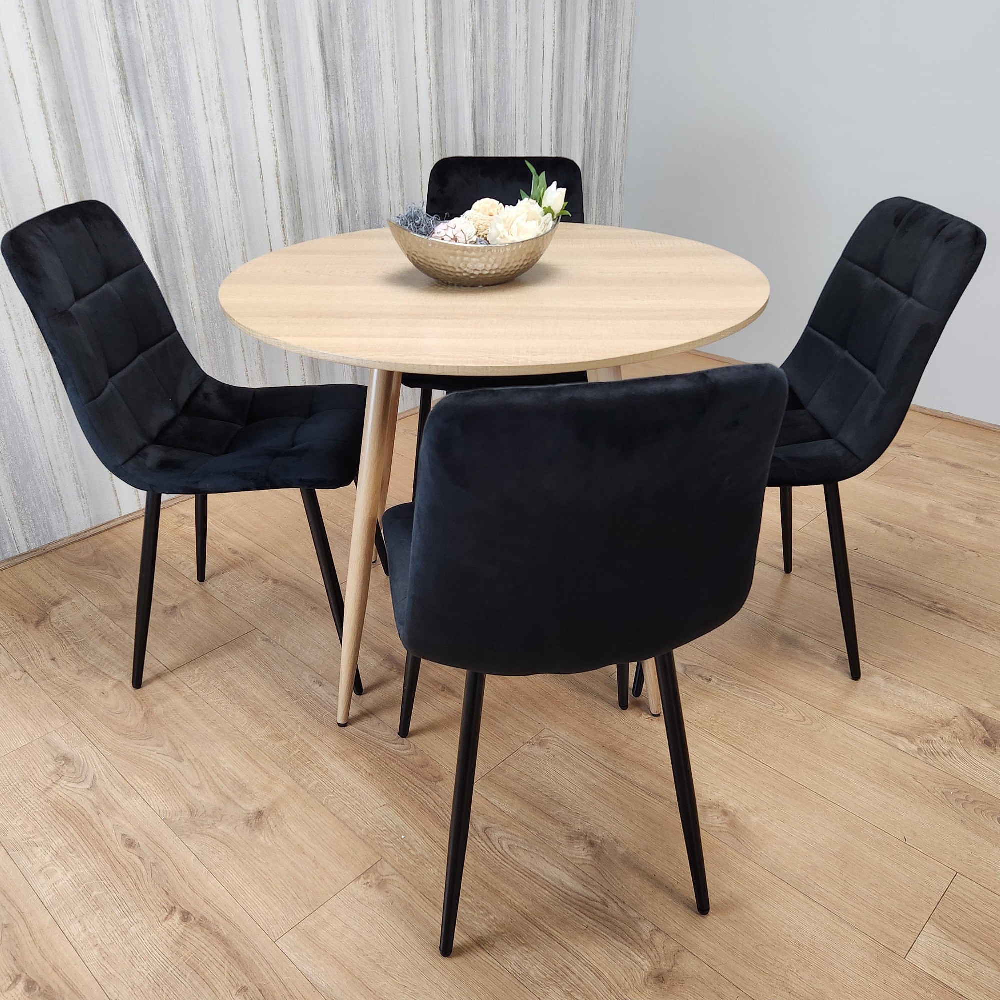 Round oak effect dining table with a smooth surface, accompanied by four black velvet tufted chairs featuring sleek black legs, set against a light wood floor and neutral wall.