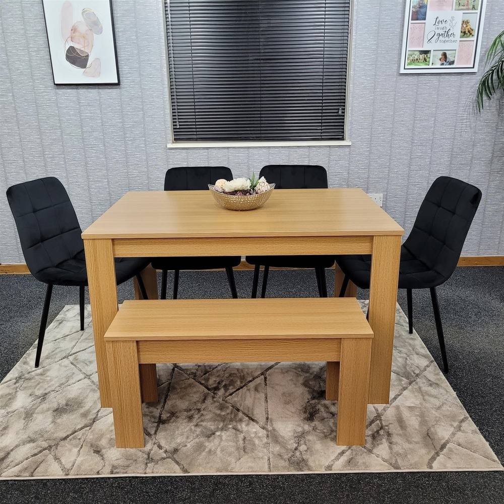 Beige wooden dining table set with four black upholstered chairs and a matching bench, placed on a patterned rug in a modern dining area.