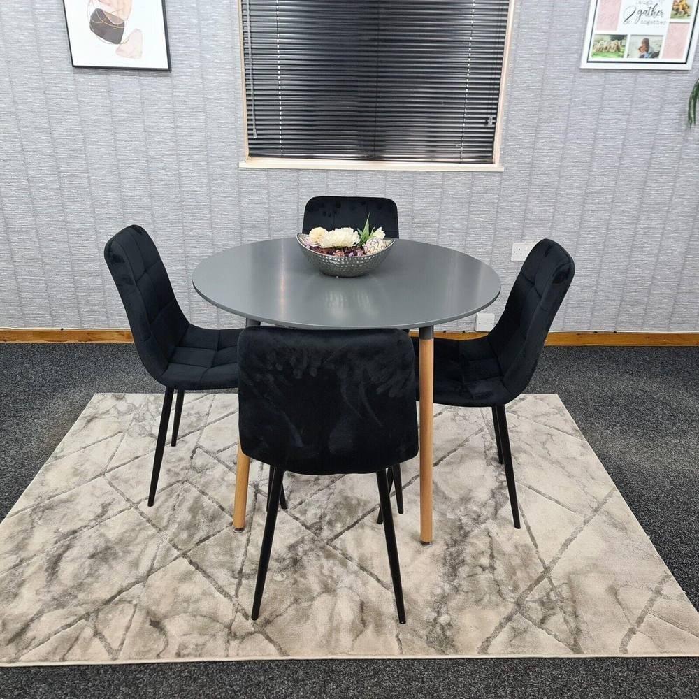 Grey round dining table set with four black velvet chairs featuring wooden legs, placed on a patterned rug in a modern dining room setting.
