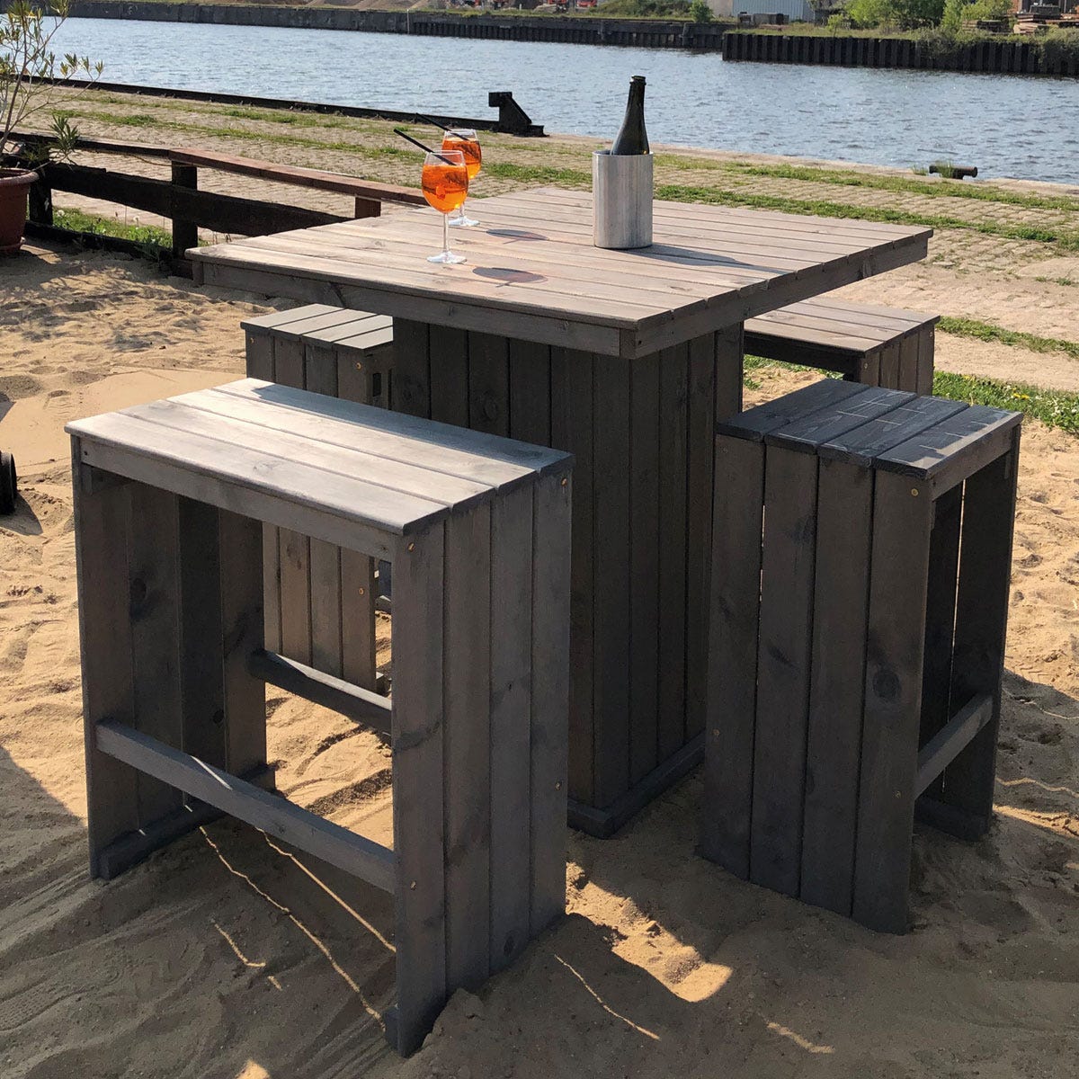 Gray wooden garden bar table with matching stools, featuring a square top and slatted design, set on sandy ground beside a body of water, with a drink and bottle on the table.