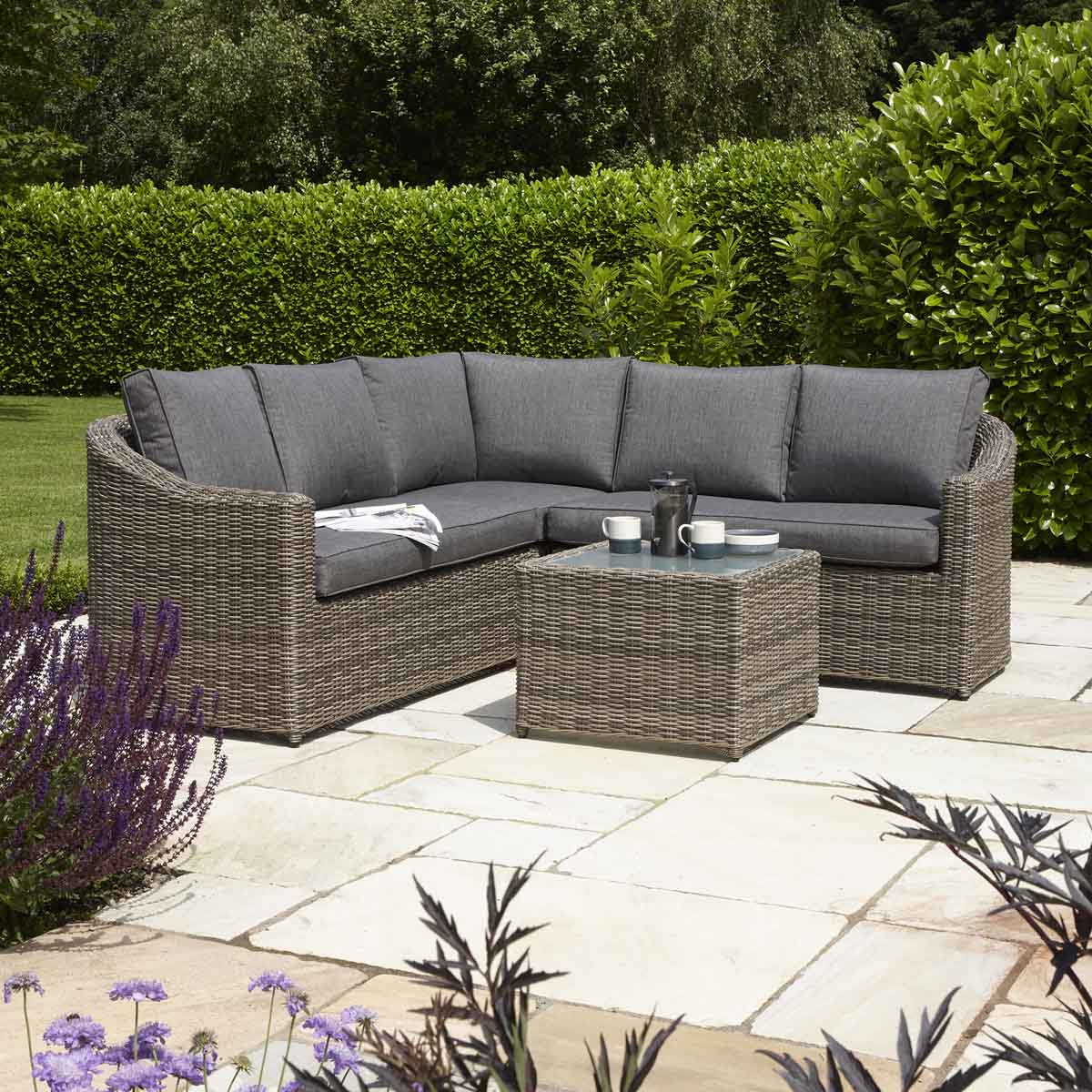 4-seater corner sofa set in natural weave rattan with light brown aluminum frame, featuring gray cushions and a matching square coffee table on a stone patio surrounded by greenery.