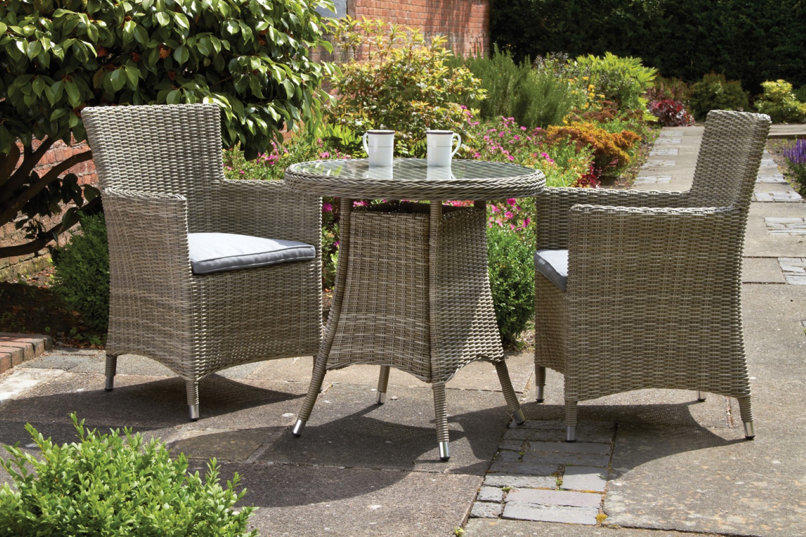 Stylish 2-seater bistro set featuring a 70cm round glass table and two grey wicker carver chairs with soft cushions, set in a garden with vibrant greenery.
