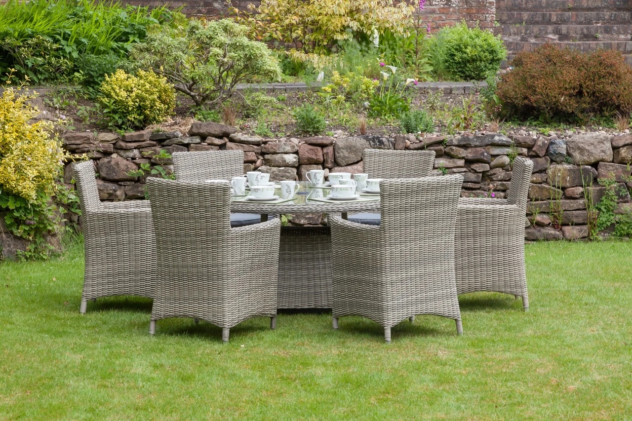 Round 140cm dining table with six grey wicker carver chairs, each featuring cushions, set on a green lawn against a stone wall and garden backdrop.