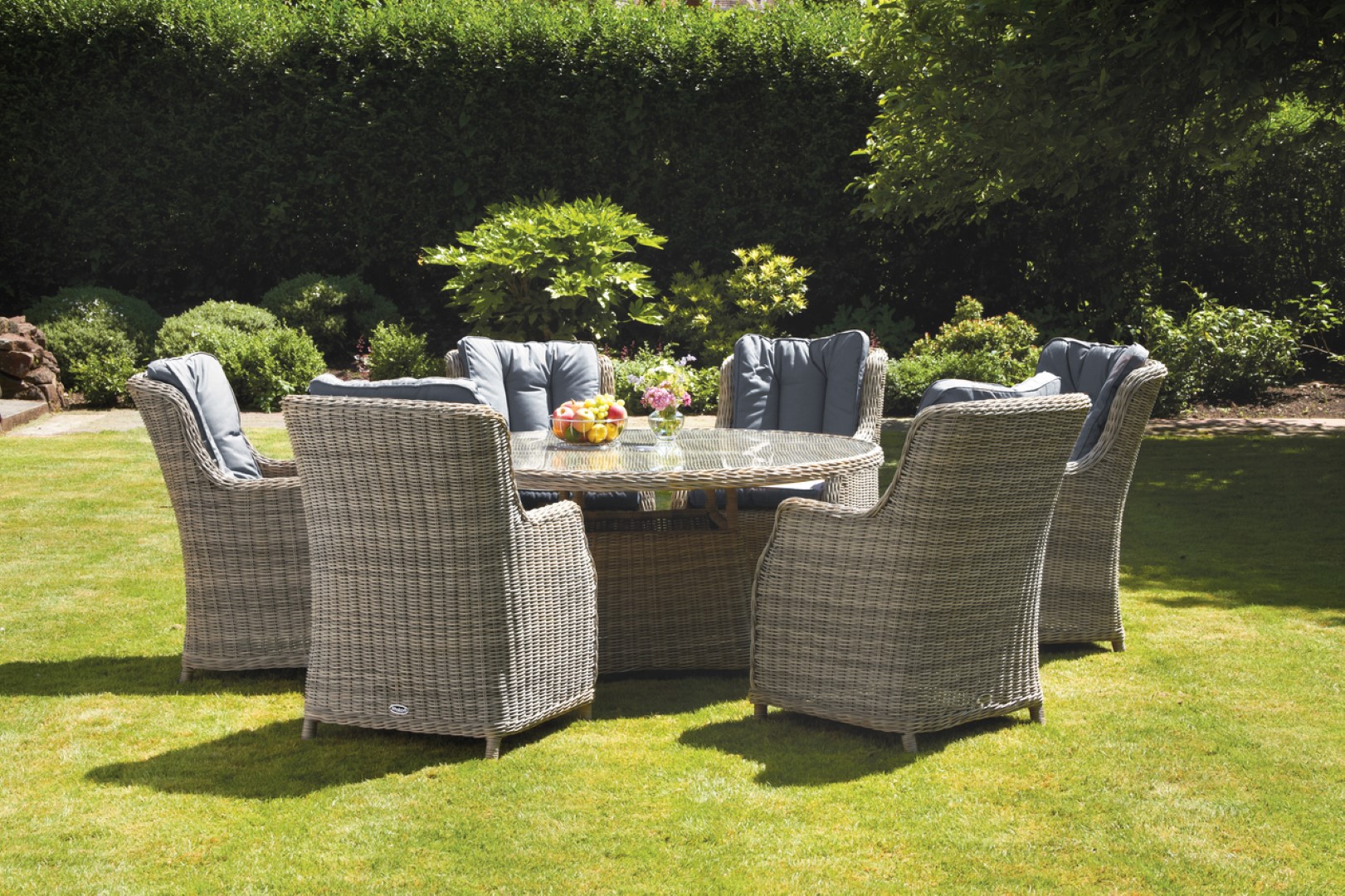 Royalcraft Wentworth 6-seat round dining set featuring cream-colored highback chairs with soft cushions, a woven rattan base, and a glass tabletop, set on a lush green lawn.