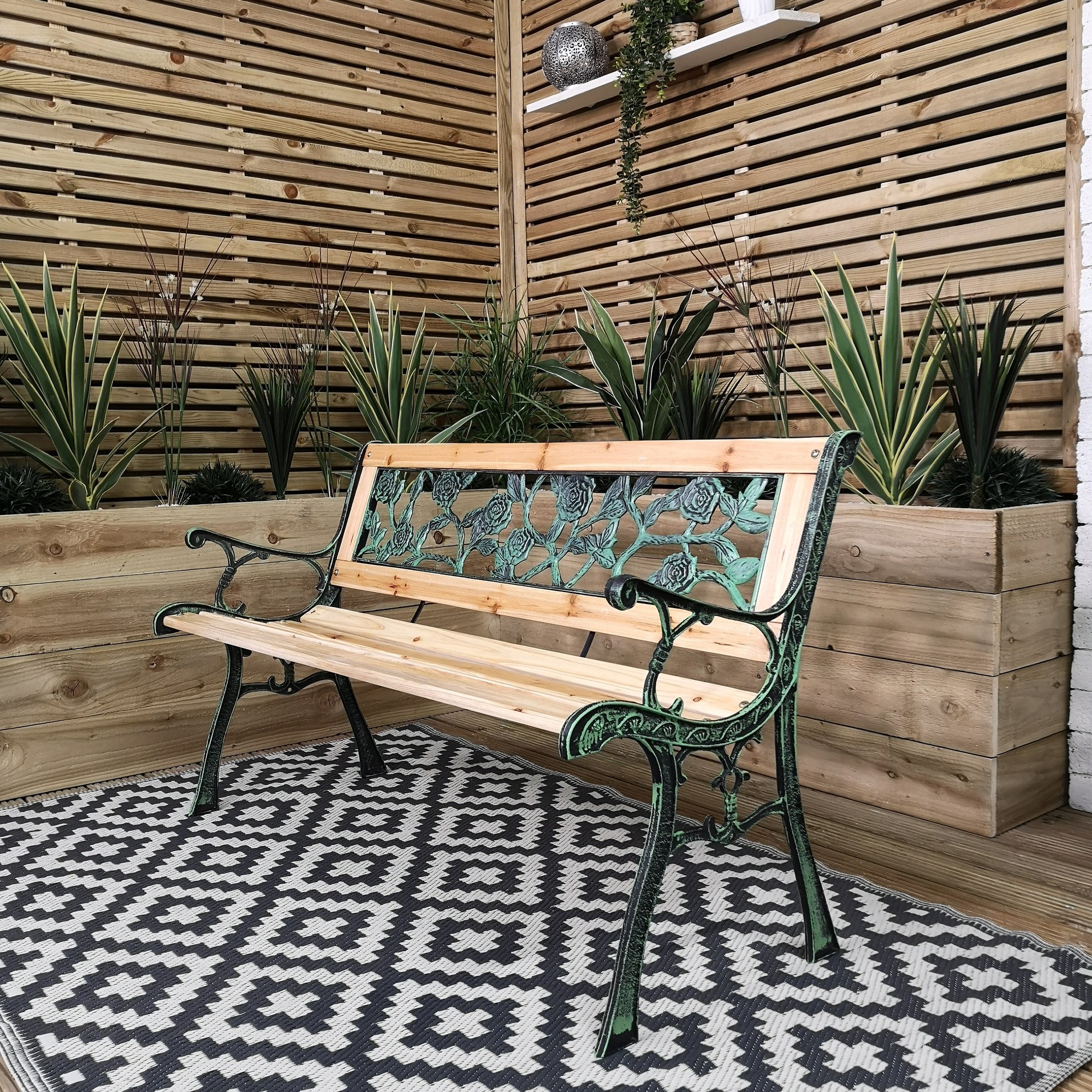 A two-seater garden bench featuring a wooden seat and ornate green cast iron arms with a rose design, set against a backdrop of wooden slats and potted plants.