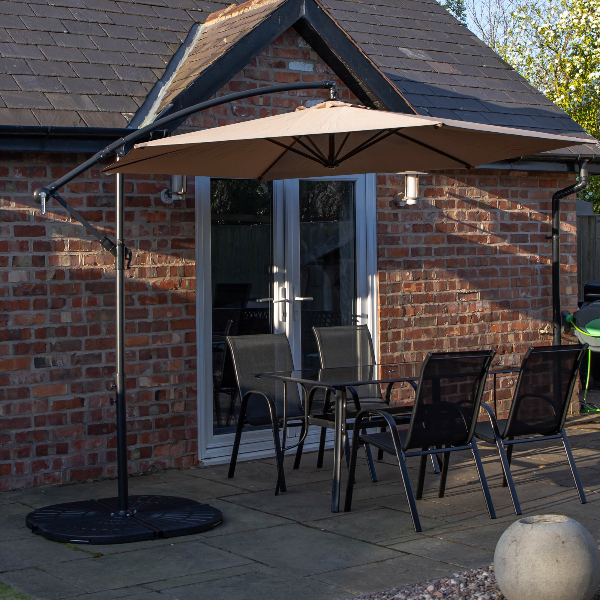 Rectangular glass top dining table with four black metal chairs, accompanied by a beige banana parasol and sturdy base, set against a brick wall backdrop.