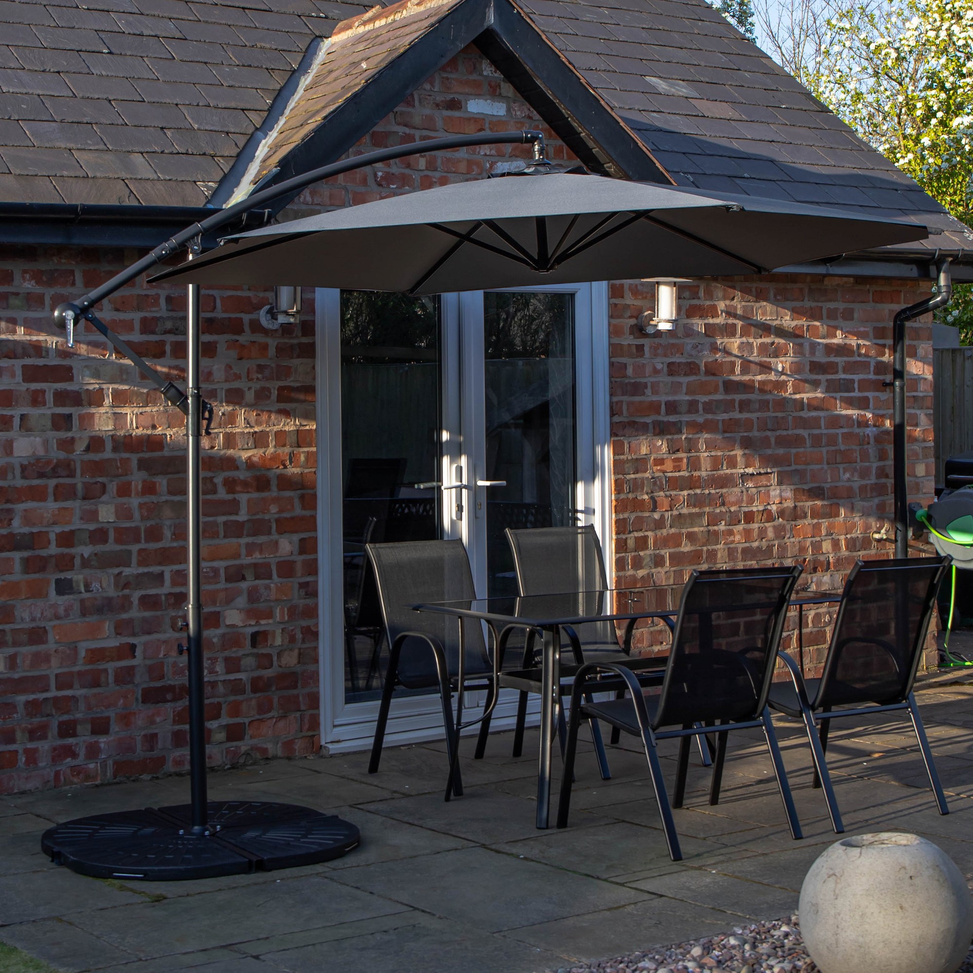 Rectangular glass top table with four black chairs, accompanied by a grey banana parasol and base, set on a stone patio near a brick house.