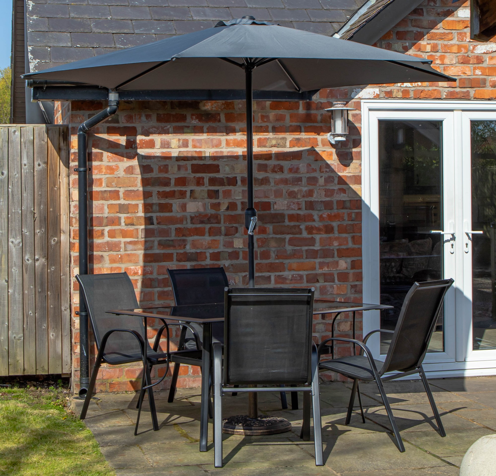 Rectangular glass top dining table with four black chairs, featuring a grey Havana parasol and base set, situated on a stone patio near a brick wall.
