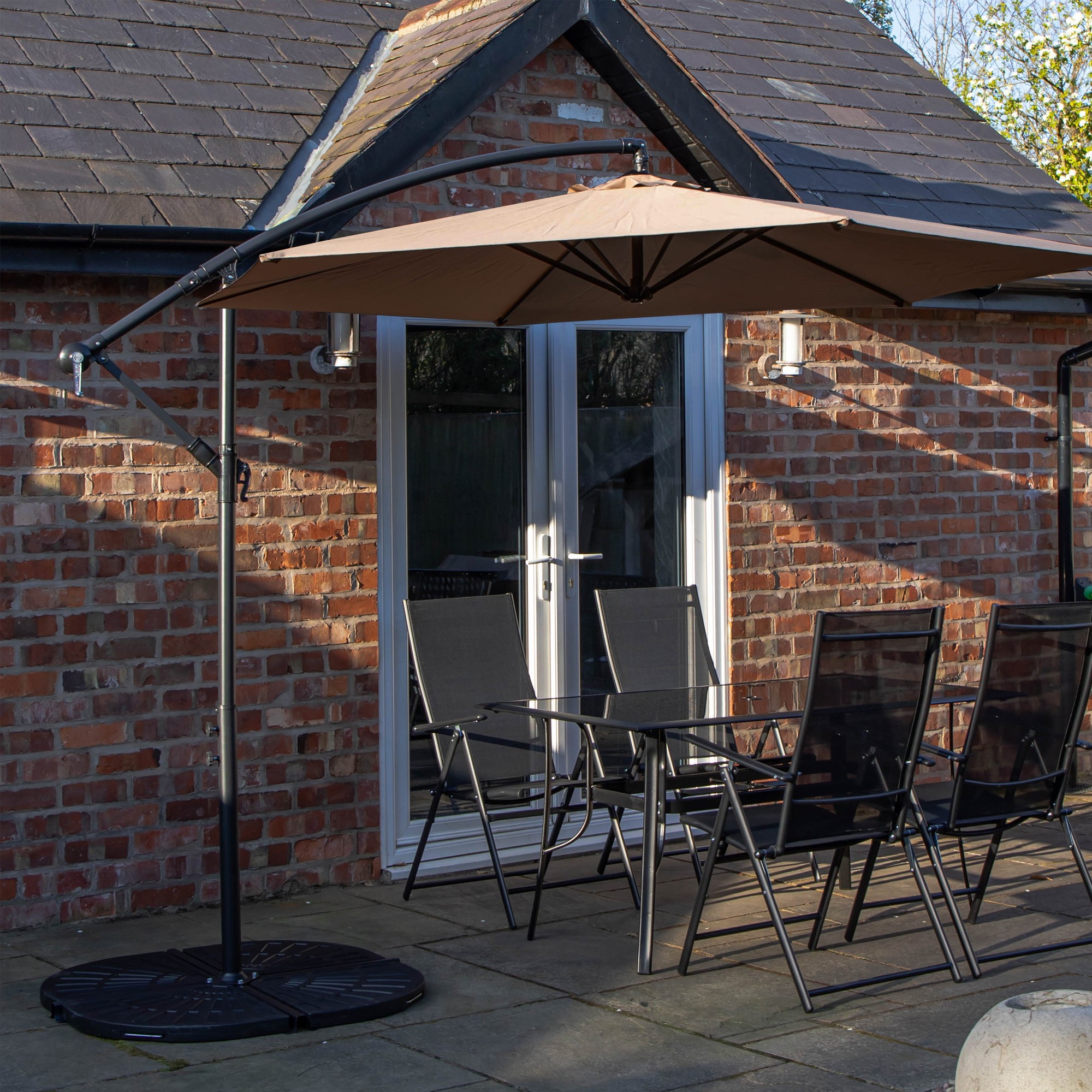 Rectangular glass top table with four black chairs, accompanied by a beige banana parasol and base, set against a brick wall and patio.