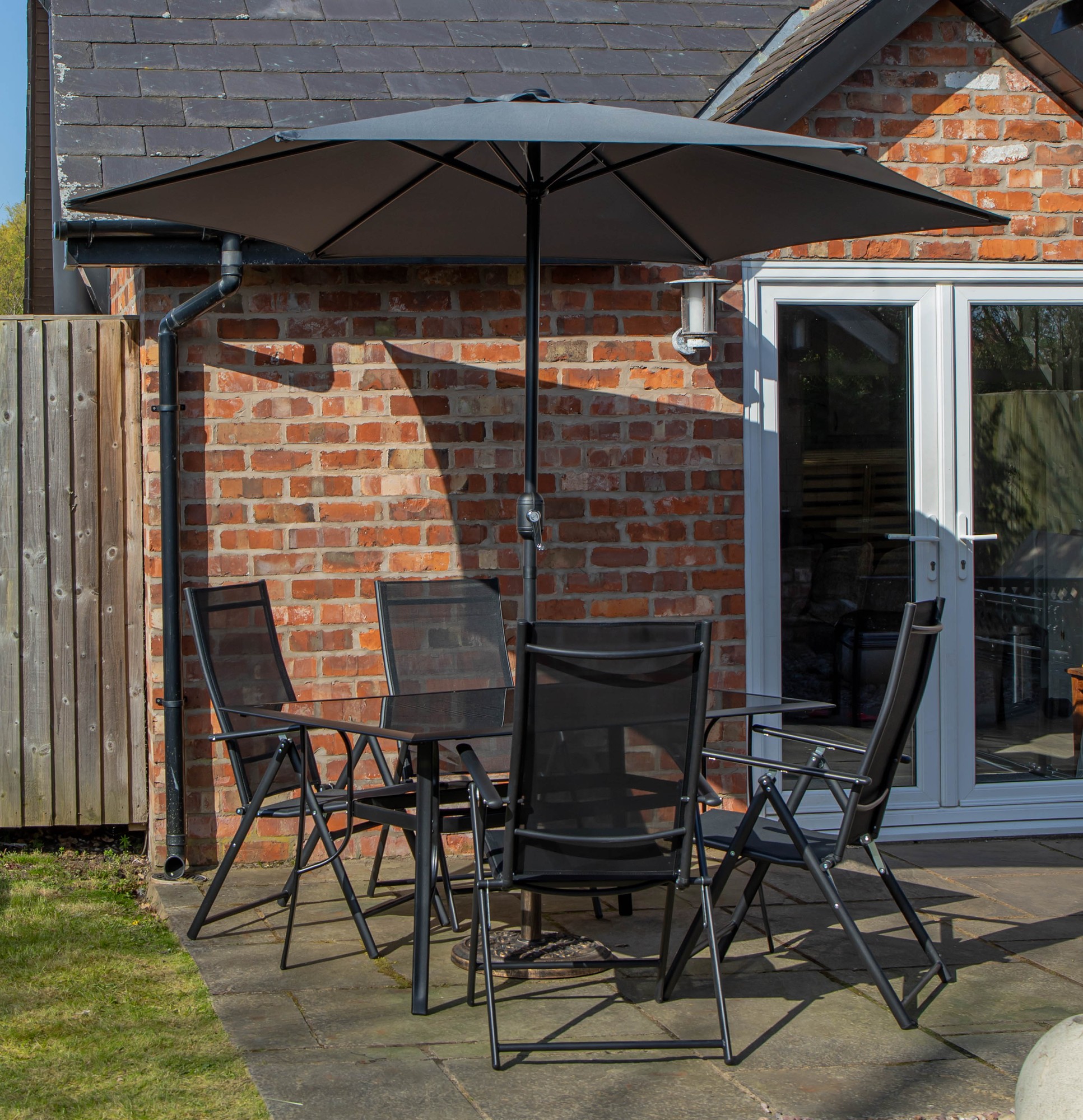 Rectangular glass top dining table with four black mesh chairs, accompanied by a grey Havana parasol and base, set against a brick wall and patio.