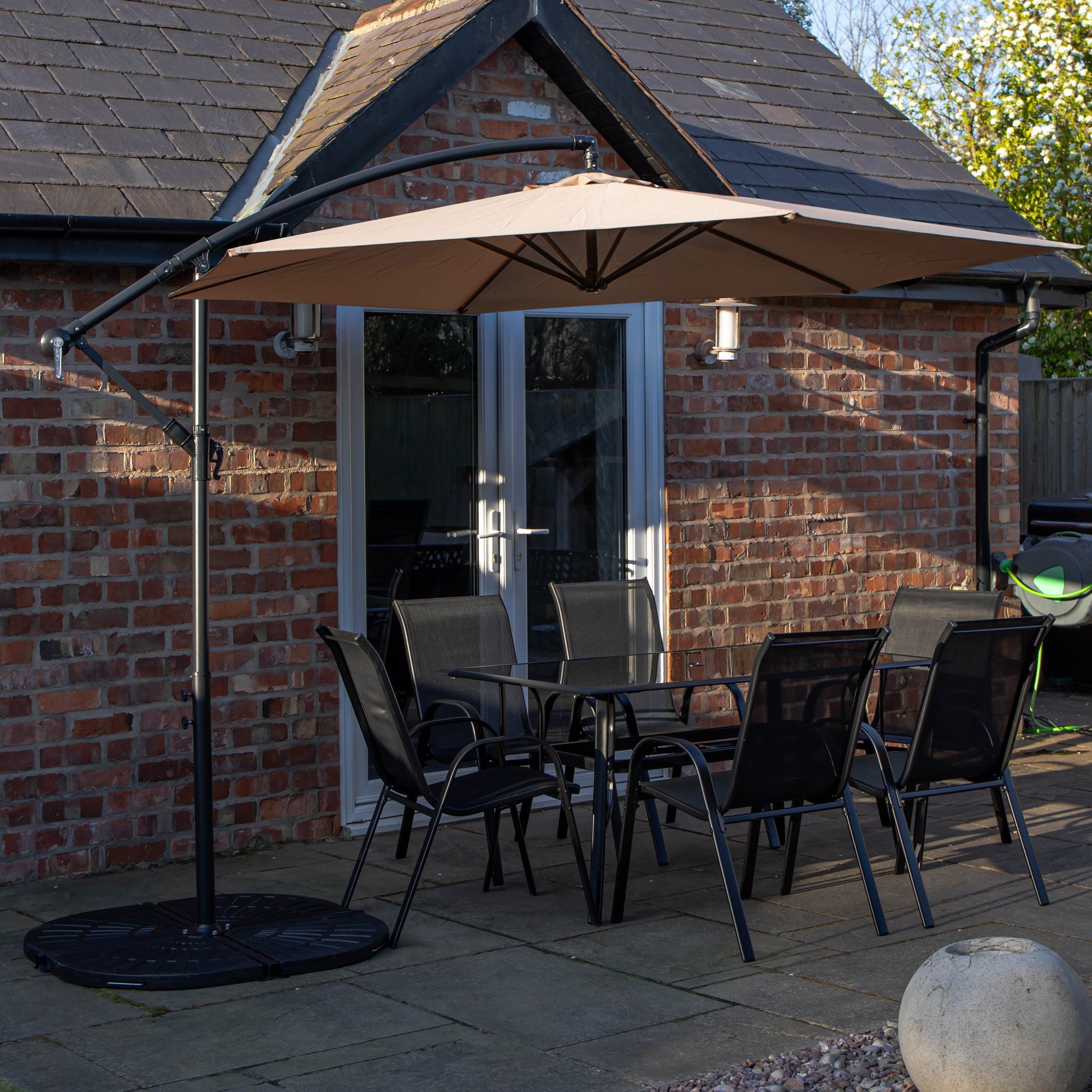 Rectangular glass top dining table with six black metal chairs, accompanied by a beige banana parasol and base, set against a brick wall backdrop.