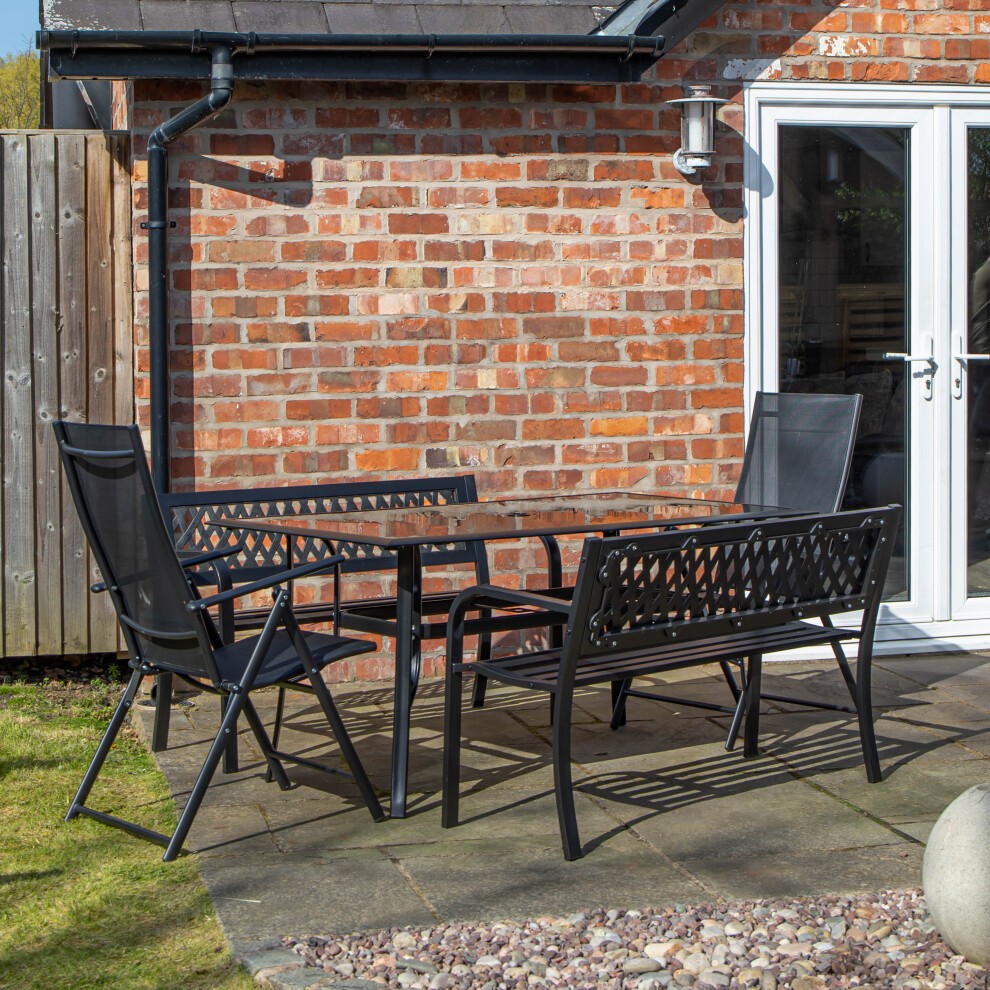 Rectangular glass top dining table with black metal frame, accompanied by two matching benches and chairs, set against a brick wall and grassy area.