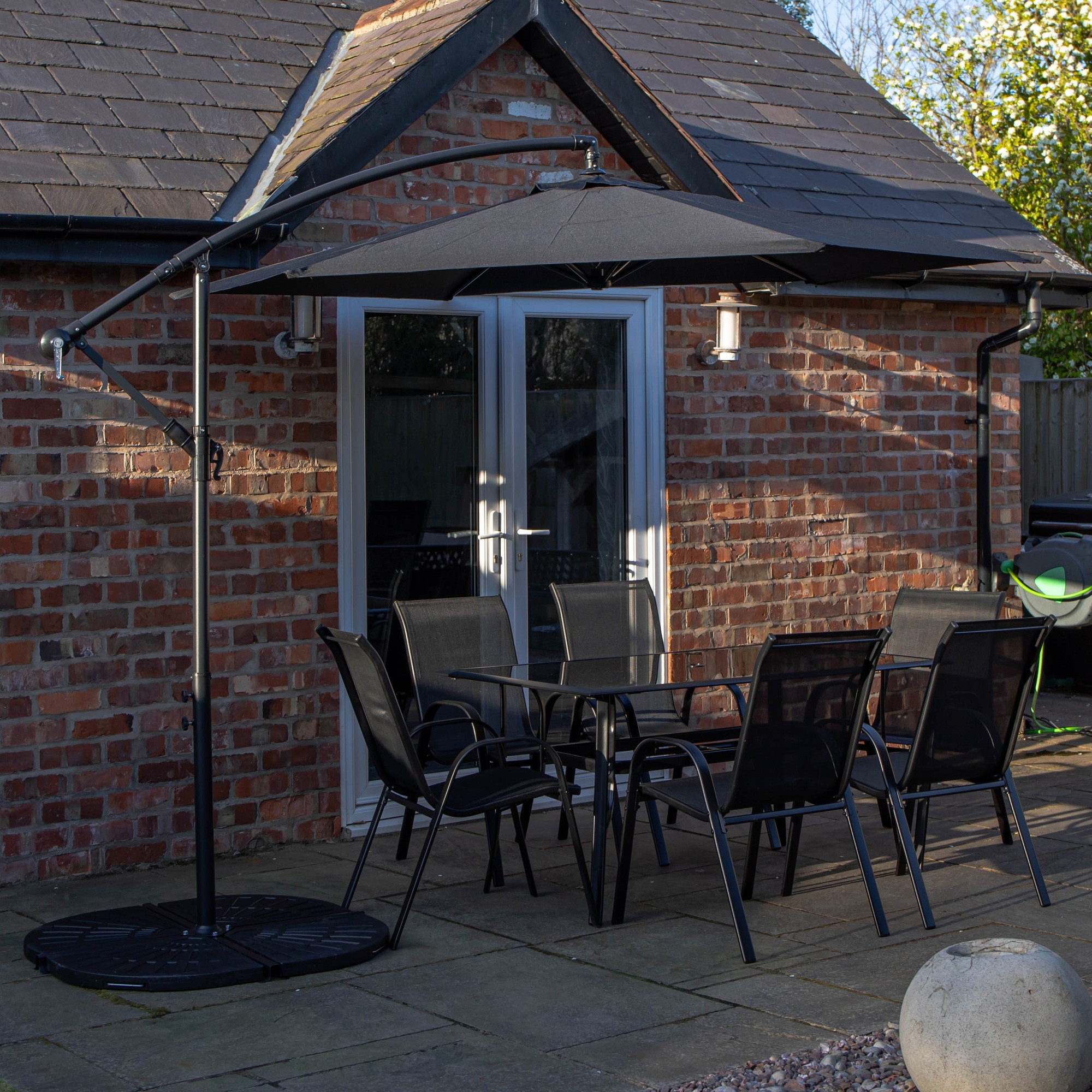 Rectangular glass-top dining table with six black chairs, accompanied by a black banana parasol and base, set against a brick wall and patio.