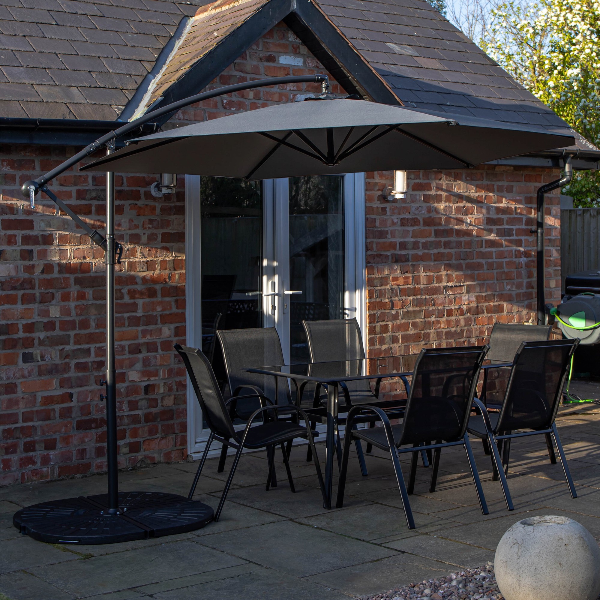 Rectangular glass top dining table with six black chairs, accompanied by a grey banana parasol and base, set on a patio with a brick wall backdrop.