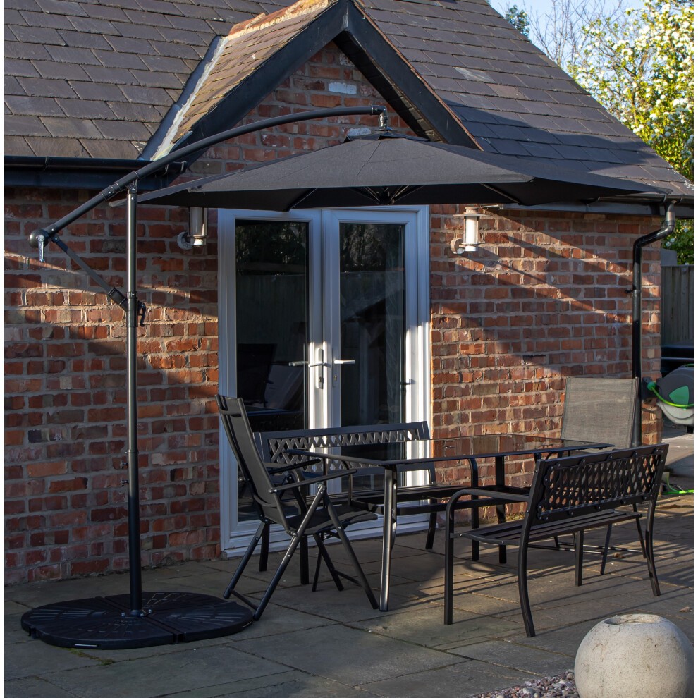 Rectangular glass top table with black metal benches and chairs, accompanied by a black banana parasol and base, set against a brick wall and patio.