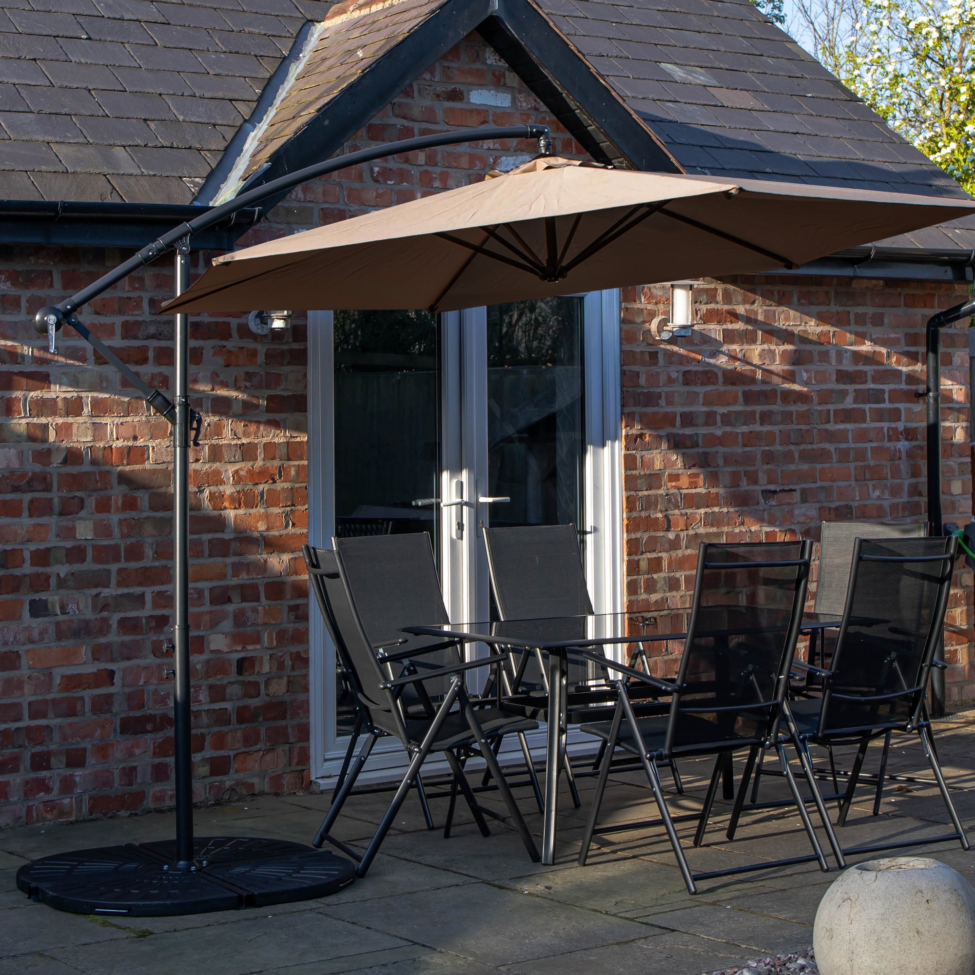 Rectangular glass top table with six black chairs, featuring a beige banana parasol and sturdy base, set on a patio with brick walls and a view of double doors.