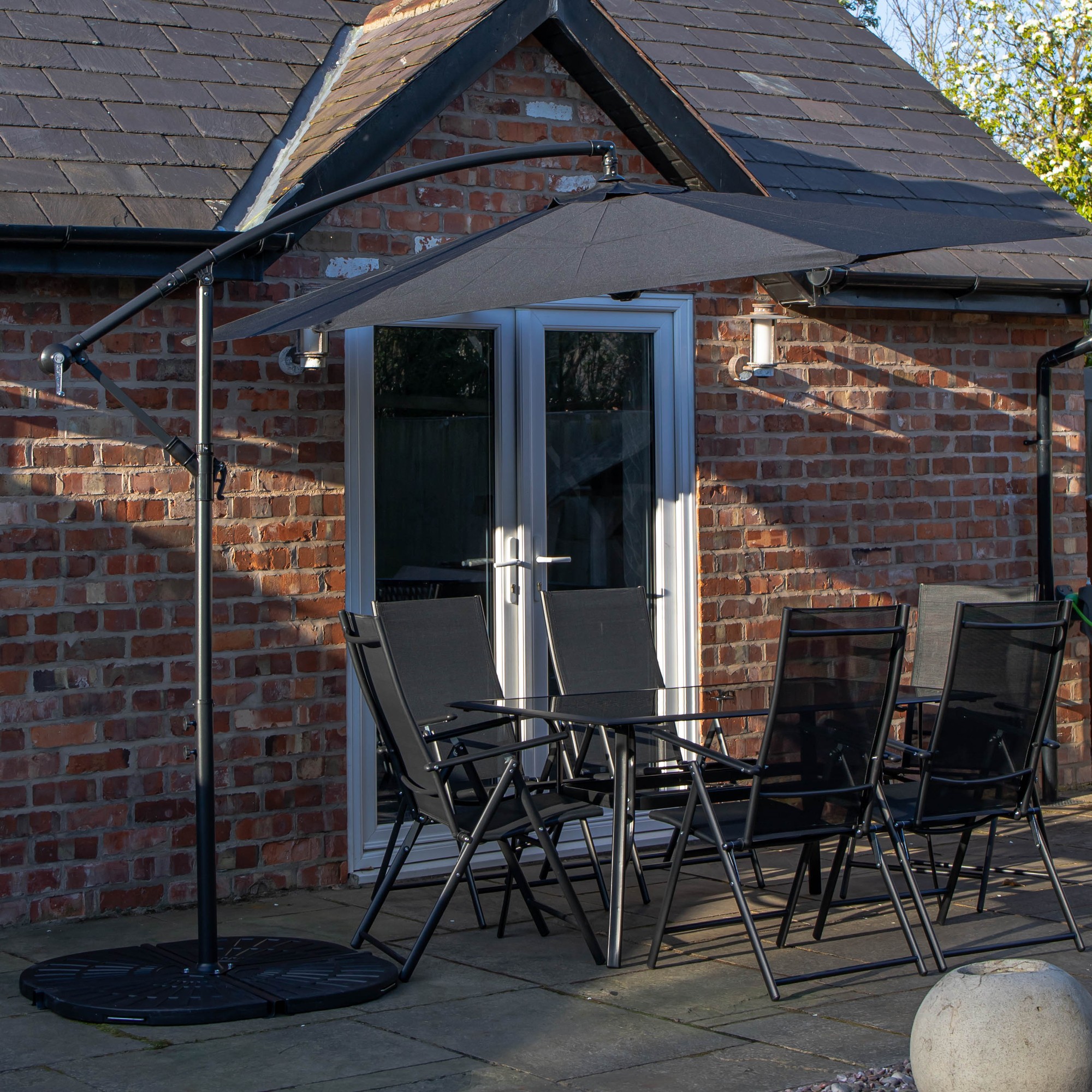 Rectangular glass top table surrounded by six black chairs, featuring a black banana parasol with a sturdy base, set against a brick wall backdrop.
