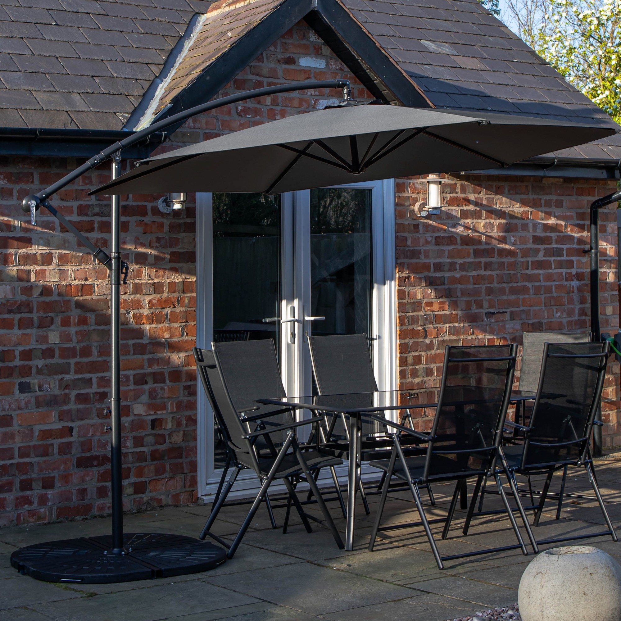 Rectangular glass top table surrounded by six black chairs, topped with a grey banana parasol and base, set on a patio with a brick wall backdrop.