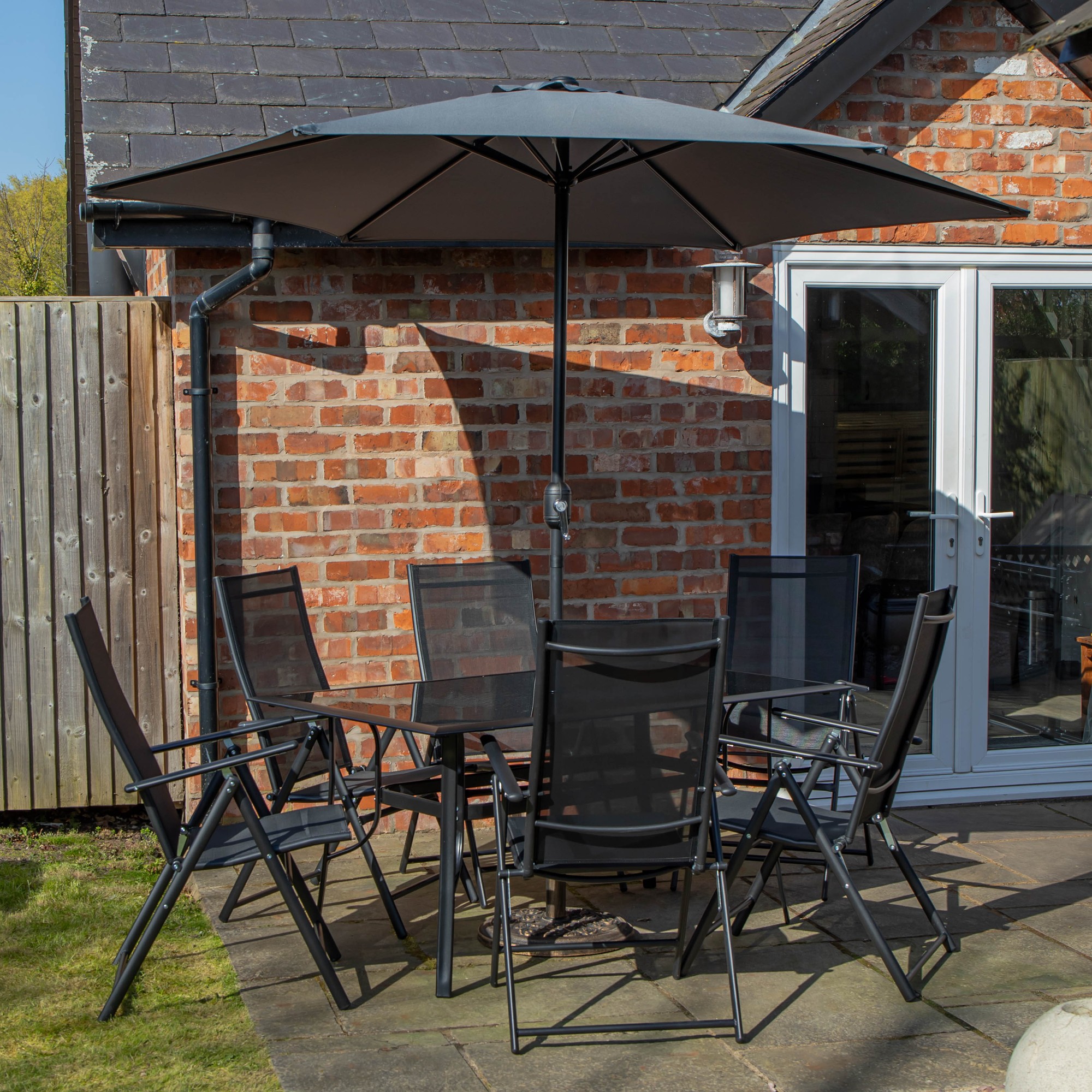 Rectangular glass top dining table with six black chairs, accompanied by a grey Havana parasol and base, set in a brick patio area.