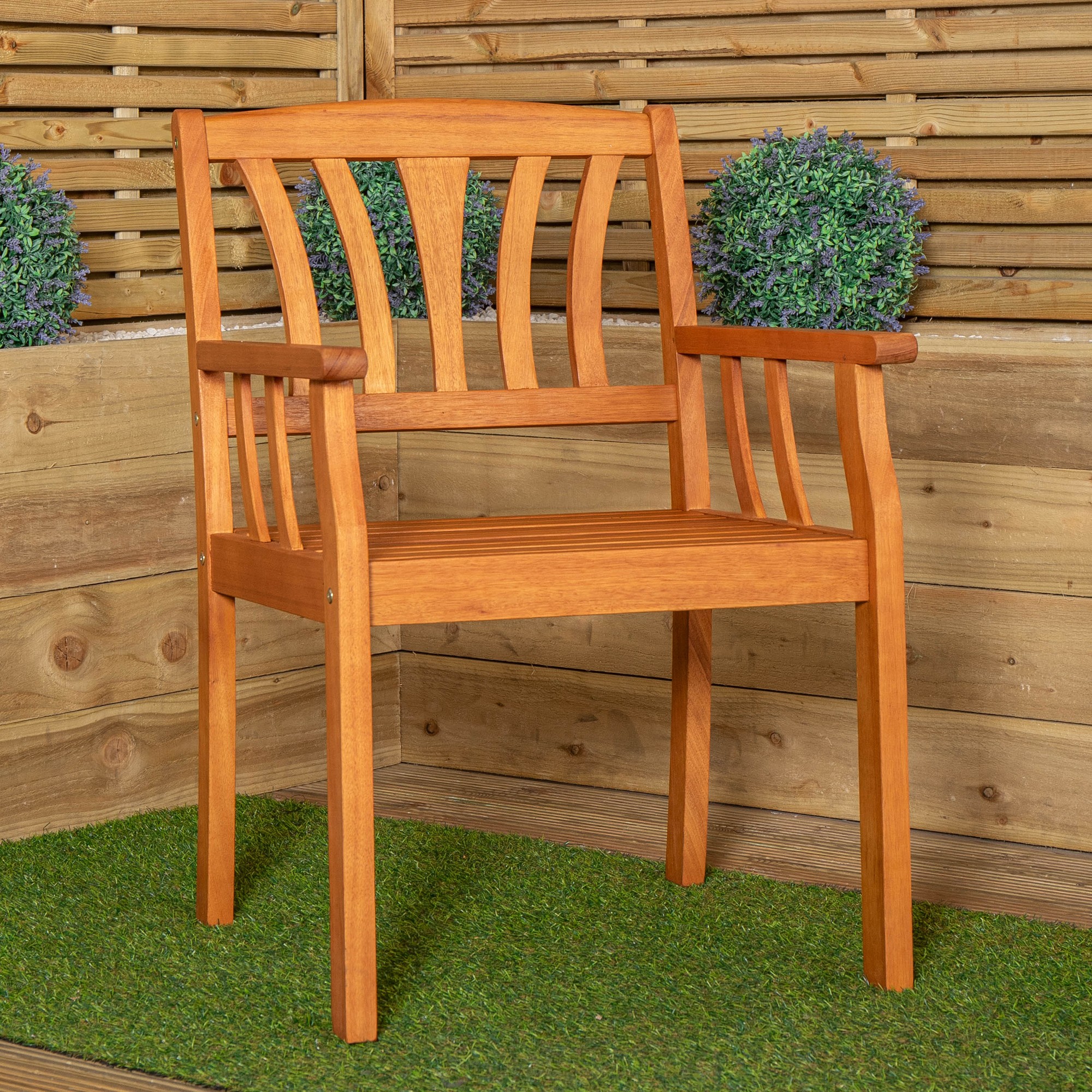 Brown wooden garden dining chair with slatted backrest and armrests, featuring a sturdy design, placed on green grass against a wooden backdrop with potted greenery.