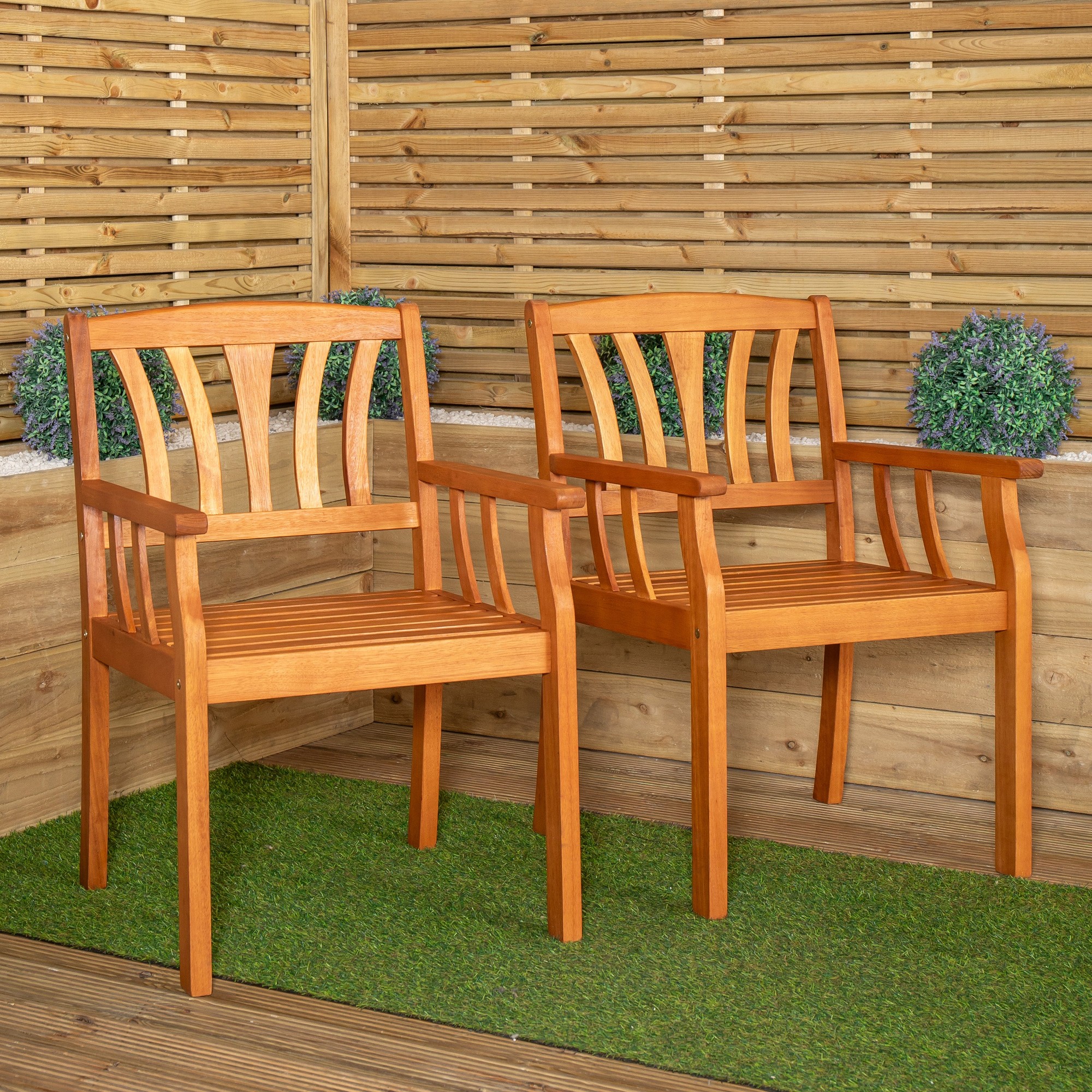 Set of 2 brown wooden garden dining chairs featuring slatted backs and seats, with armrests, placed on green grass-like flooring against a wooden backdrop.