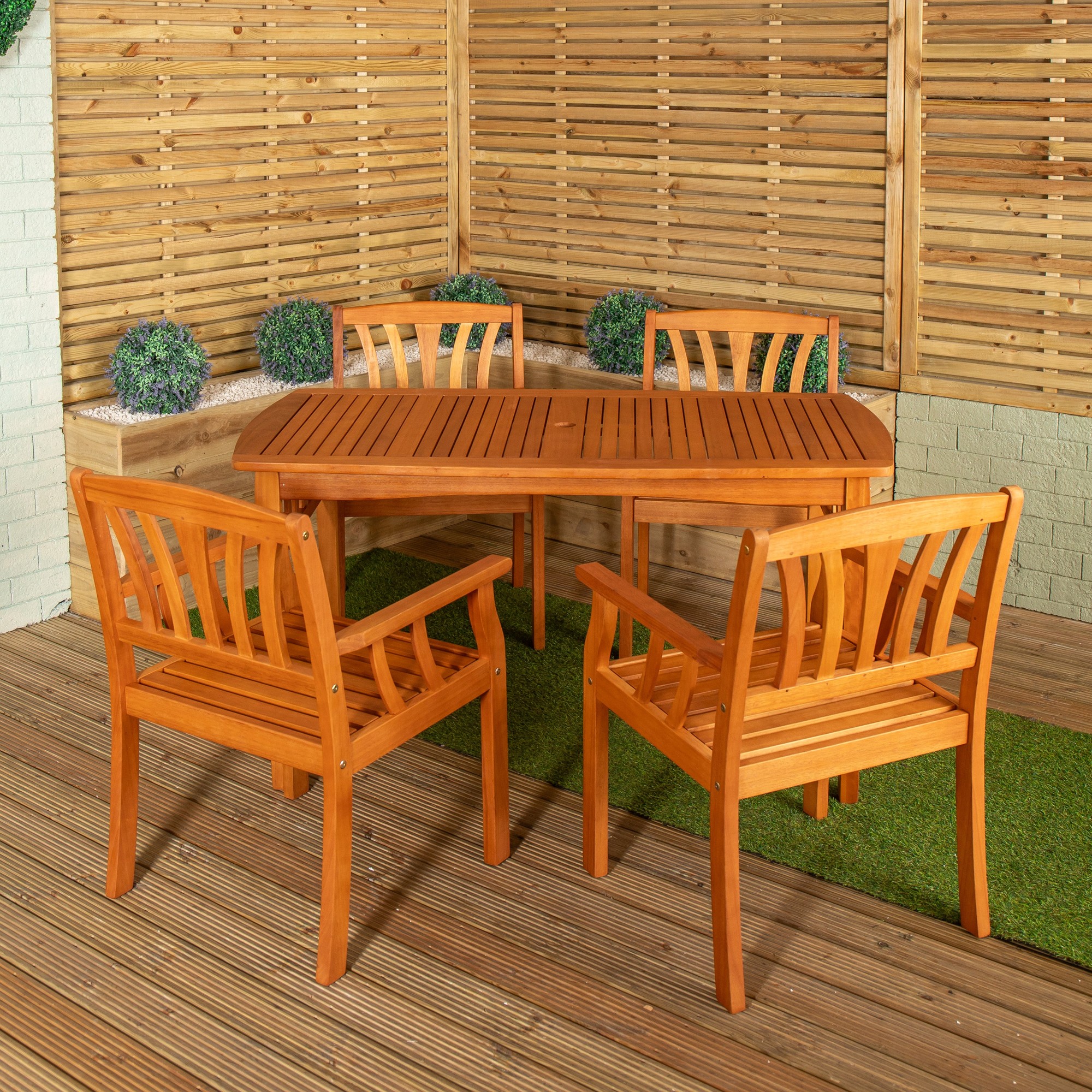 Rectangular wooden dining table with slatted top and four matching chairs, all in a warm honey finish, set on a wooden deck with green grass accents and a wooden fence backdrop.