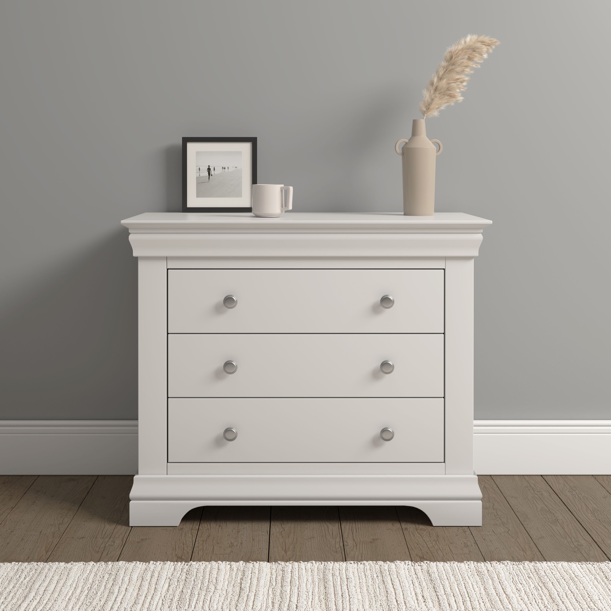 White wooden 3-drawer chest with silver knobs, featuring a classic design, a smooth top surface, and a subtle decorative molding, set against a gray wall.