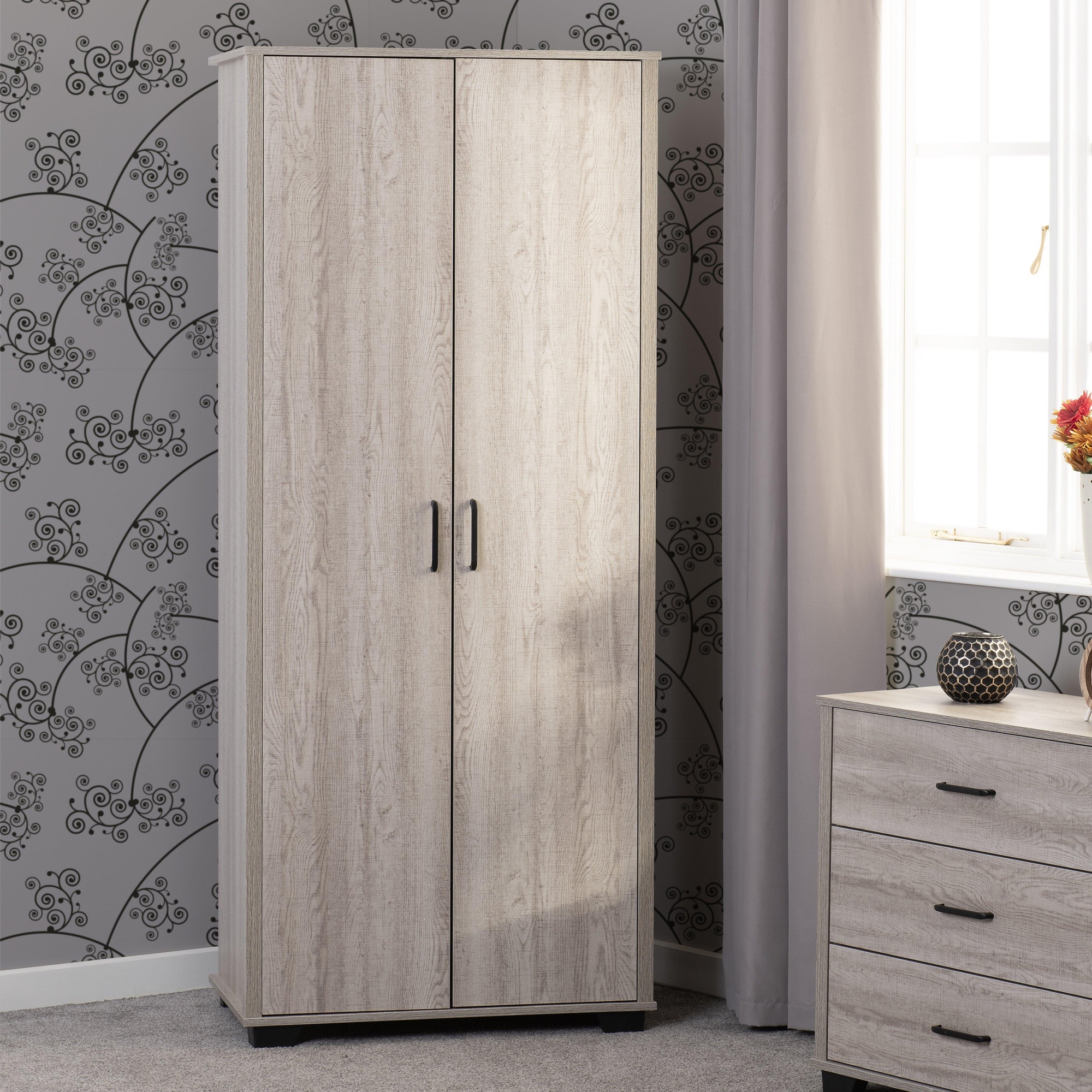 Light oak effect wardrobe with two doors, featuring sleek black handles, set against a patterned grey wall, complemented by a nearby dresser and natural light from a window.