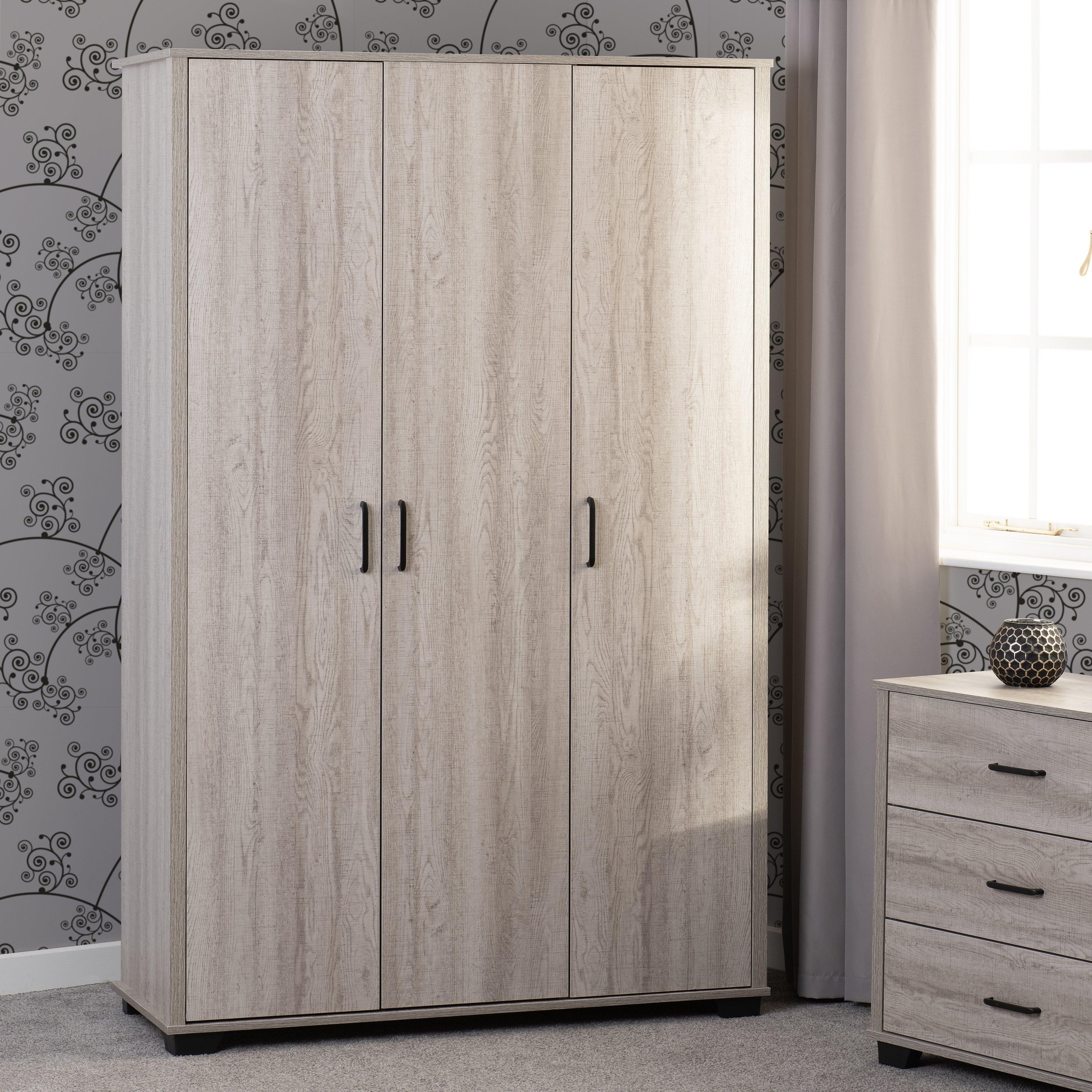 Light oak effect wardrobe with three doors, featuring a textured finish and sleek black handles, set against a patterned wall and accompanied by a matching bedside cabinet.
