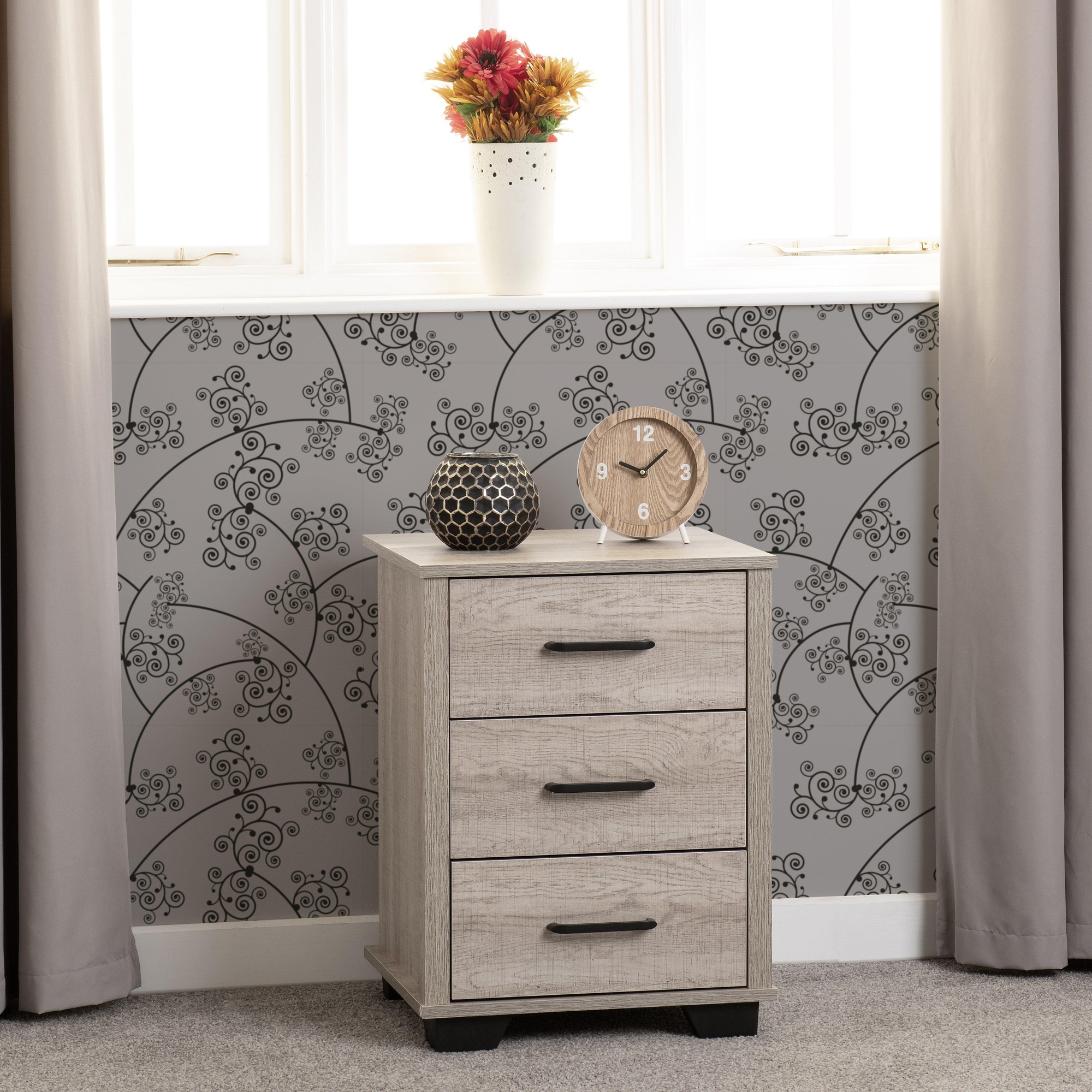 Light oak effect bedside table with three drawers, black handles, and a sturdy base, styled with a clock, decorative vase, and geometric decor against a patterned wall.