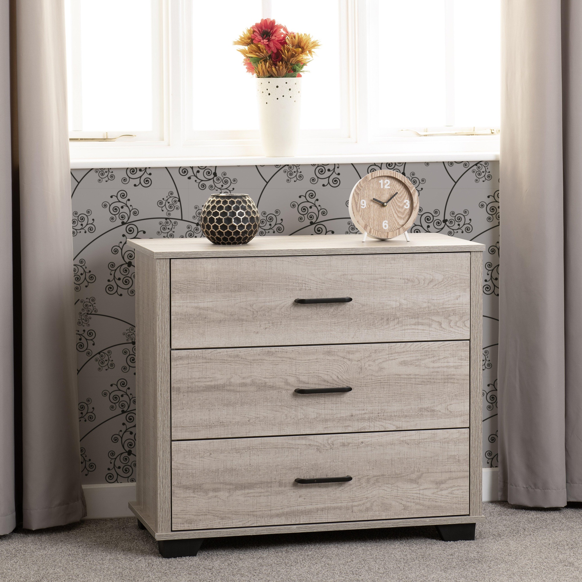 Light oak effect 3-drawer chest with a brown finish, featuring sleek black handles and a modern design, placed against a patterned wall with decorative items on top.