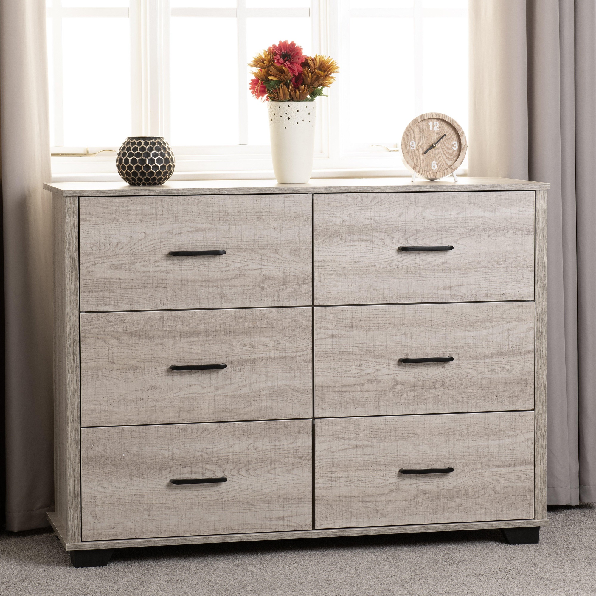 Light oak effect 6-drawer chest with a smooth finish, featuring black handles, set against a neutral backdrop with decorative items on top.