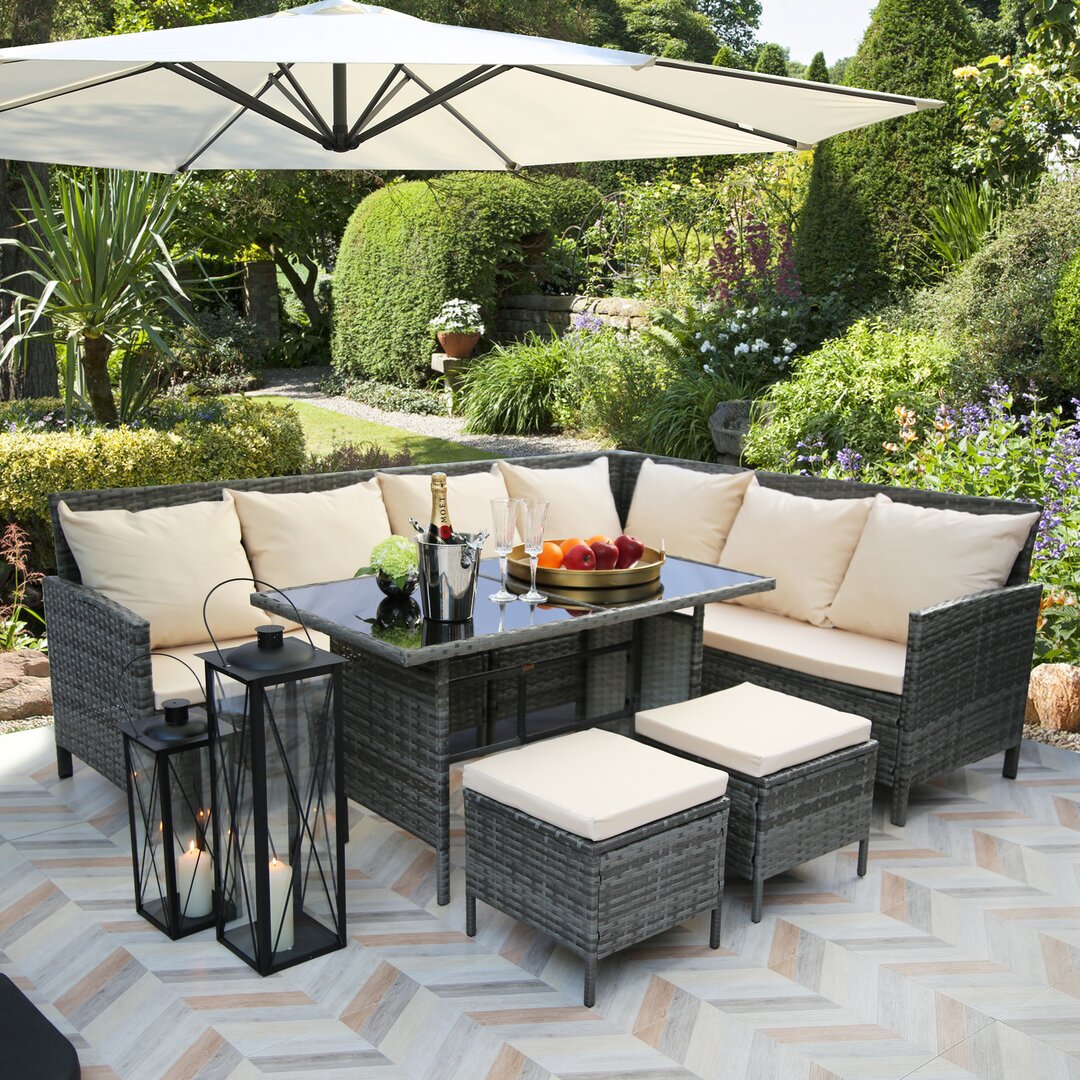 Grey rattan corner sofa dining set for eight, featuring plush cream cushions, a glass-topped table, two matching stools, and a rain cover, set in a lush garden with an umbrella.