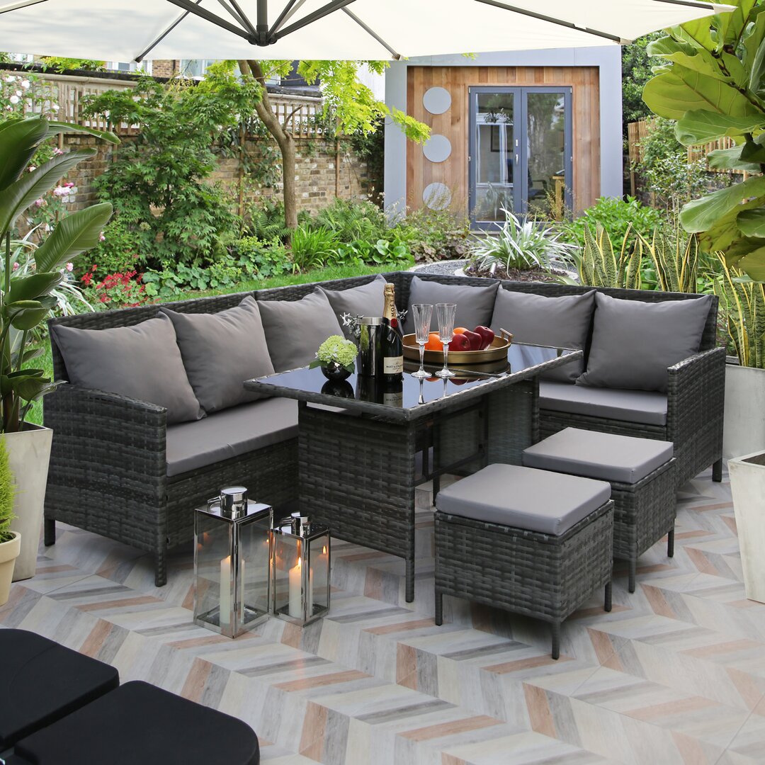 Grey rattan corner sofa dining set for eight, featuring plush grey cushions, a glass-top table, and matching stools, set in a lush garden environment.