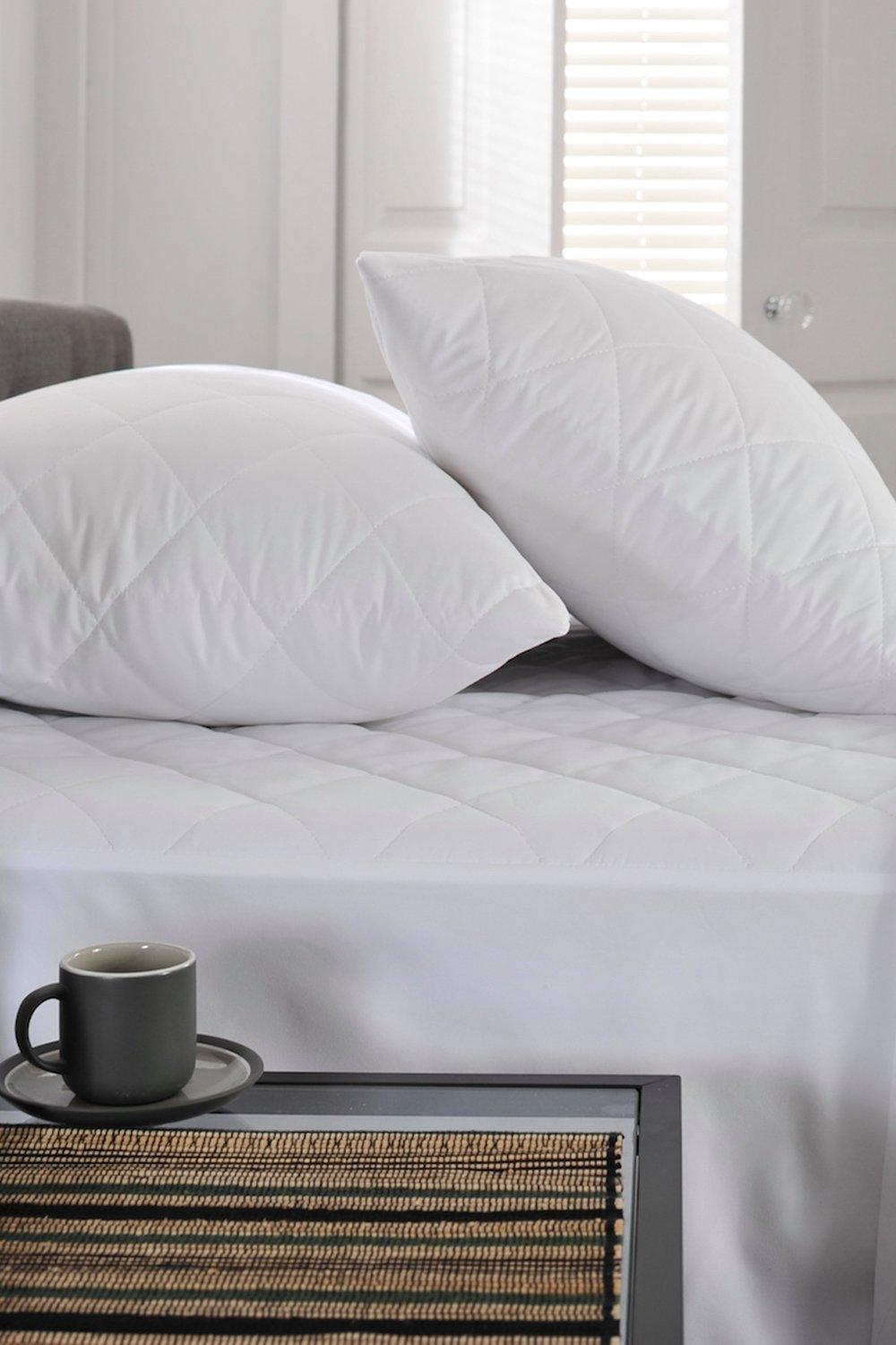 White quilted mattress protector draped over a single bed, featuring a smooth texture and diamond pattern, with two matching pillows and a dark cup on a woven side table.