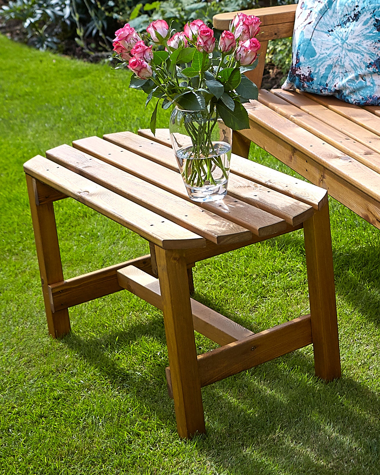 Chunky rustic wooden coffee table with a natural finish, featuring slatted top and sturdy legs, placed on green grass beside a vase of pink roses.