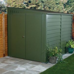 Green metal motorbike storage shed with a sloped roof, featuring double doors and horizontal slats, set on a stone patio surrounded by plants and wooden fencing.