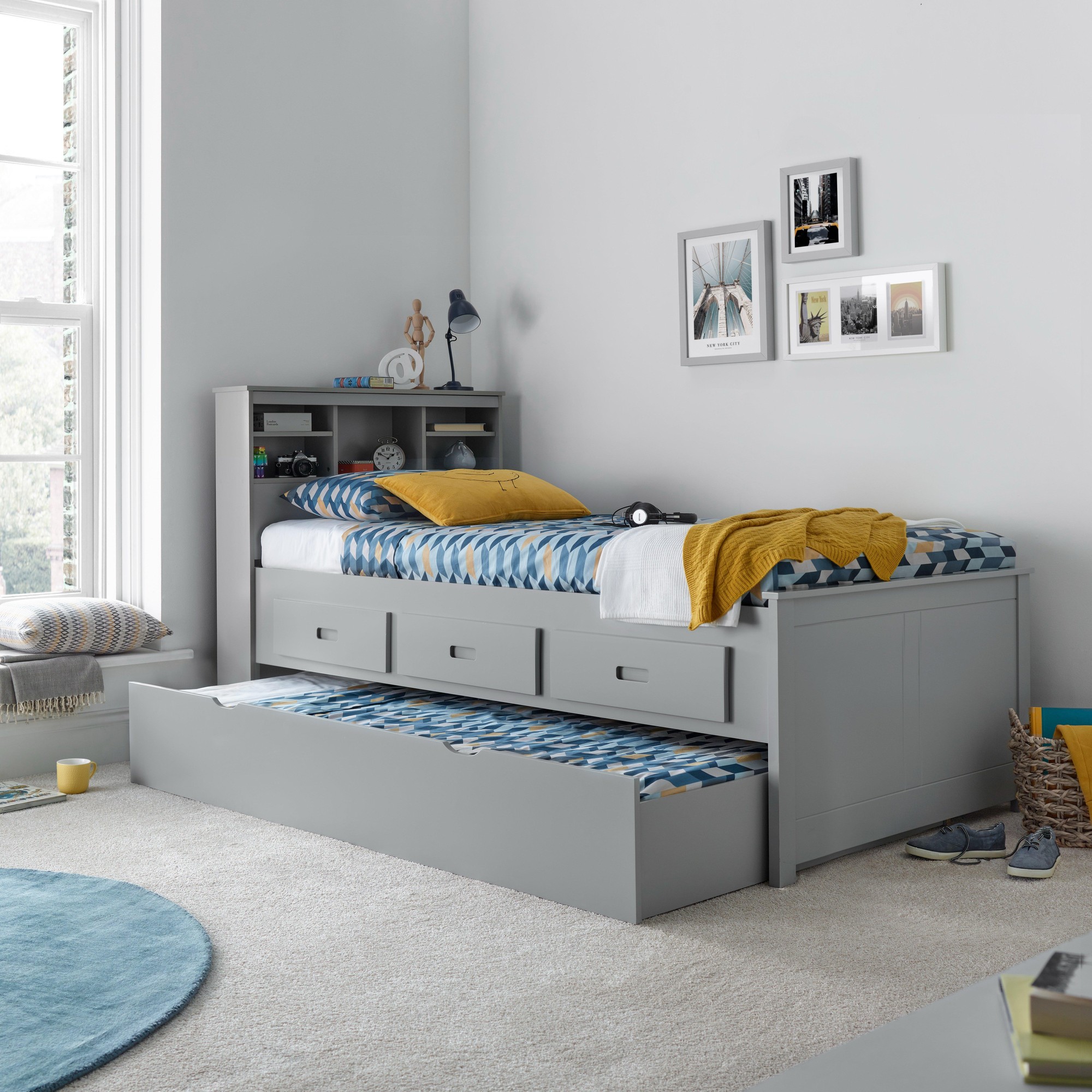 Grey guest bed with a spring mattress, featuring a sleek design, three storage drawers, and a cozy patterned bedding set, set against a light-colored room.