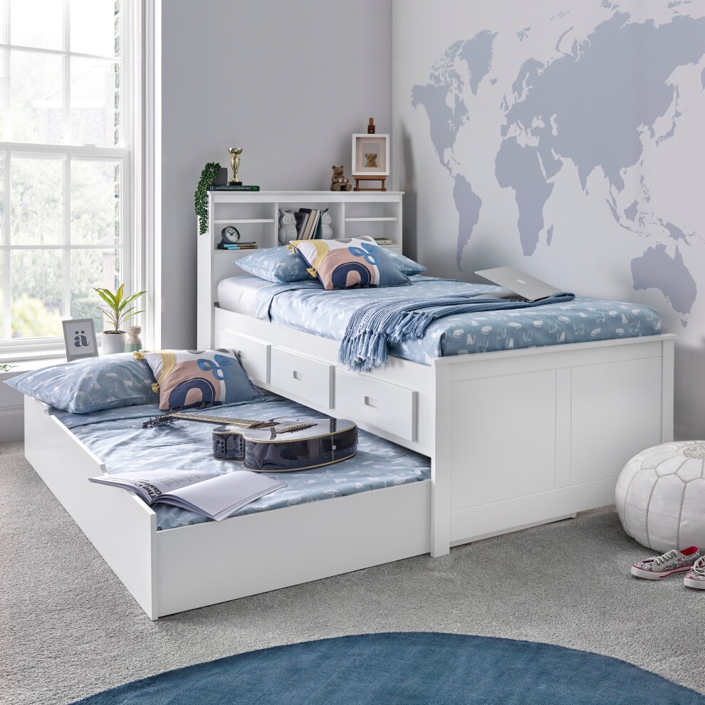 White wooden guest bed with a spring mattress, featuring two drawers and a blue bedding set, positioned in a bright room with a world map wall and a cozy decor.