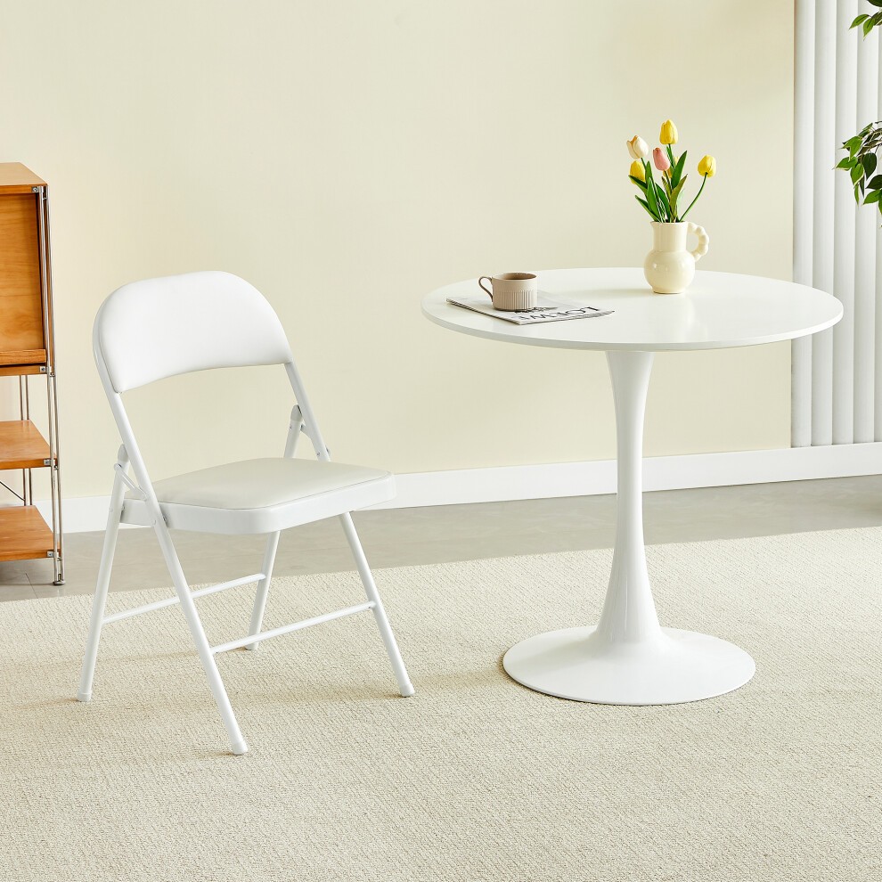 Single folding dining chair with a white metal frame and padded seat, placed next to a round white table with a vase of tulips and a cup on it, set in a light-colored room.