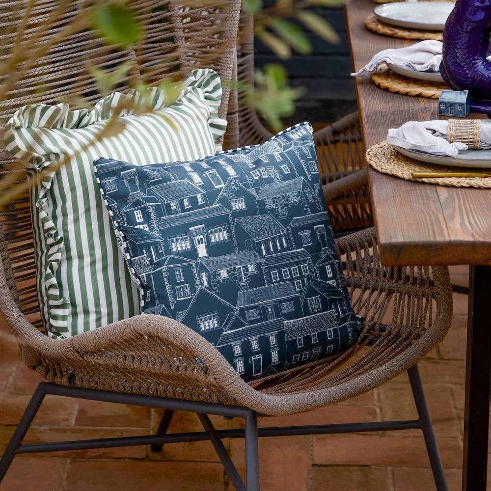 Blue outdoor cushion featuring a detailed print of houses, made from durable fabric, placed on a rattan chair alongside a striped green cushion.
